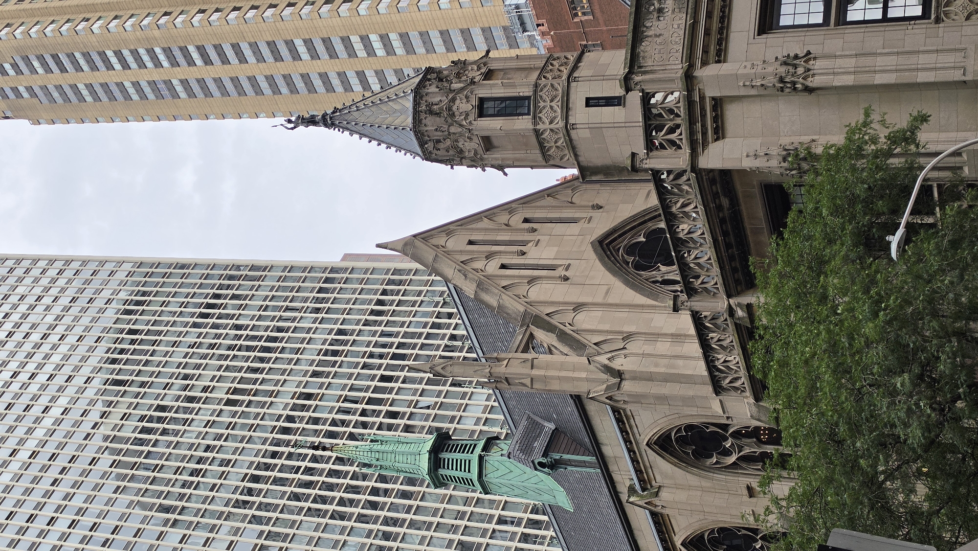 The image shows a view of the Fourth Presbyterian Church in Chicago, Illinois, United States of America. The church features Gothic-style architecture with stone spires and arched windows. A green copper spire is visible on the left side of the church, and a more elaborate stone tower with a pointed roof is on the right. The church is surrounded by modern, tall buildings with numerous windows, indicating an urban setting. Lush green trees are in the foreground, partially obscuring the lower part of the church. The sky is overcast.