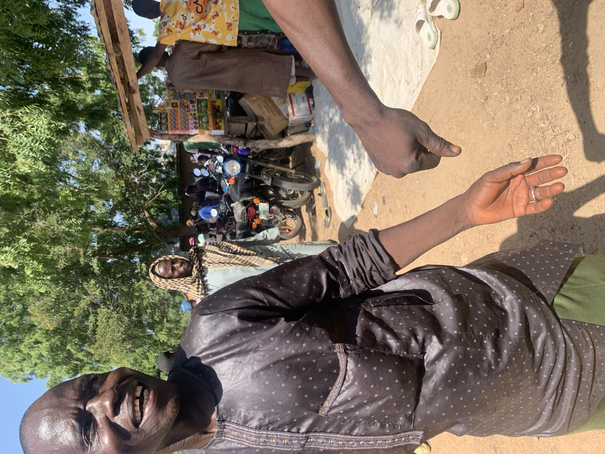 In Project, Nigeria, a man in a dark patterned tunic and green pants stands in the foreground, looking to his left and smiling broadly. His right arm is extended, and a second person's hand is visible in the lower right corner, pointing downwards with a thumb. In the background, other people are gathered around motorcycles and a market stall under a tree. The ground is sandy, and the scene is illuminated by bright sunlight.