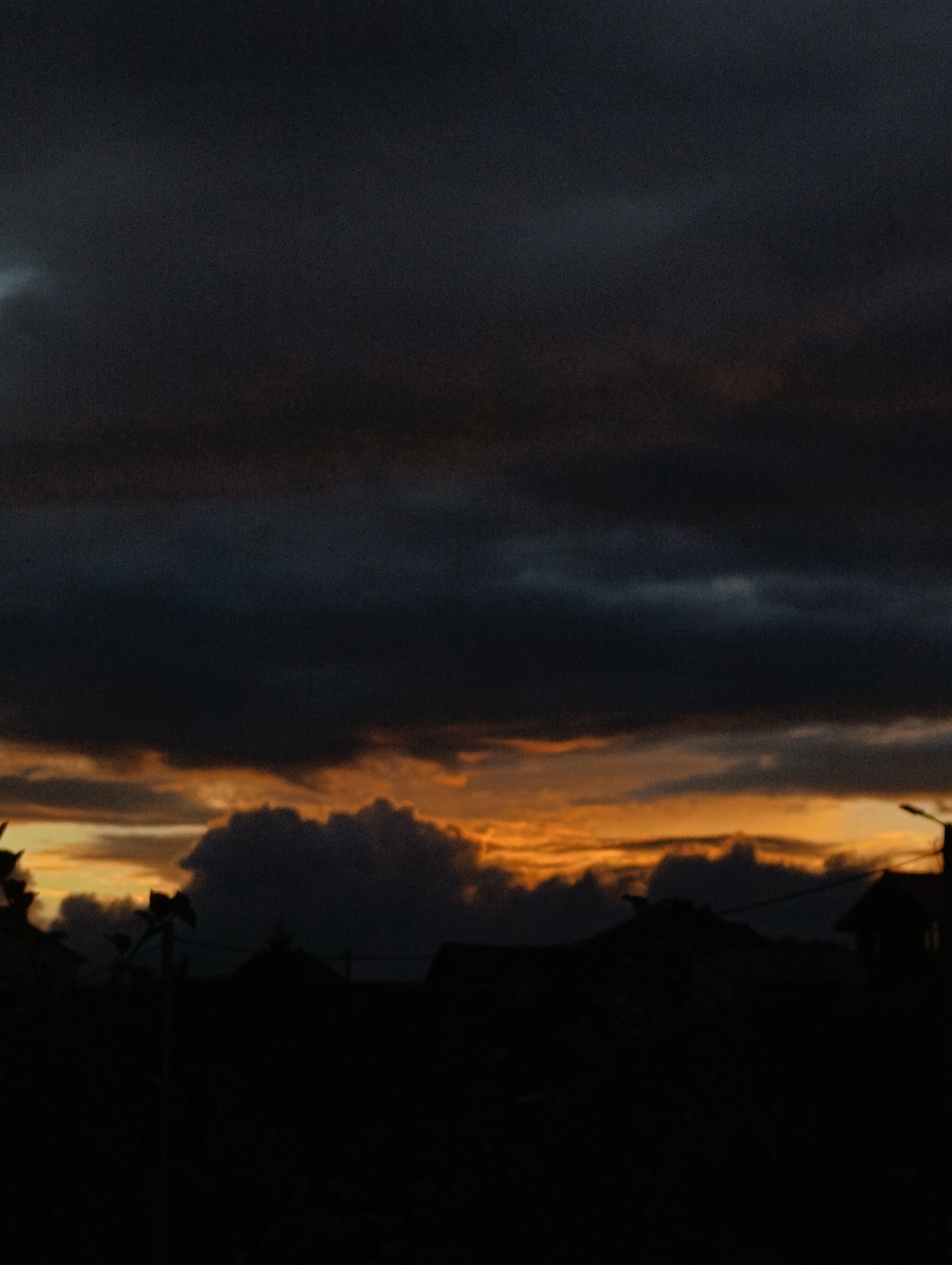 The image depicts a dark and cloudy sky with a sliver of orange light visible near the horizon, suggesting either sunrise or sunset. The lower portion of the image is obscured by the silhouettes of buildings and vegetation, making specific identification of structures or individuals impossible. The overall scene is characteristic of an urban or suburban landscape at dusk or dawn, with no discernible details that would pinpoint a location within Minsk, Belarus.