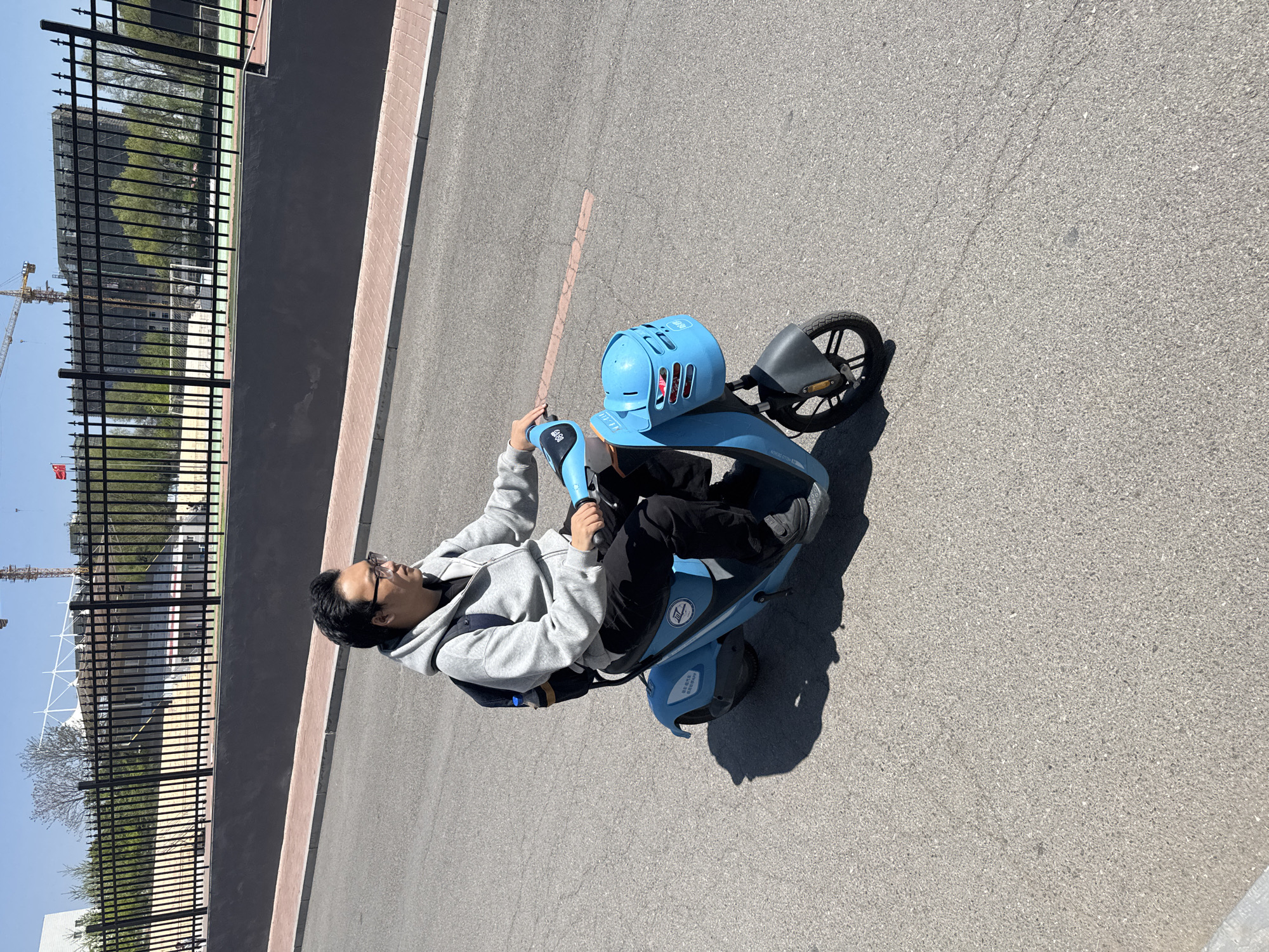 The image shows a man riding a light blue, shared electric scooter on a paved road in Zhongtun, China. The man is wearing glasses, a grey hooded sweatshirt, and black pants and shoes. He is looking towards the left side of the frame. A black metal fence runs along the left edge of the road, behind which are trees, buildings, and a construction crane. The sky is clear and blue. The road surface appears to be asphalt. The scooter has a blue basket or helmet carrier on the handlebars and a smaller wheel at the back. The overall scene suggests an urban or suburban environment on a sunny day.