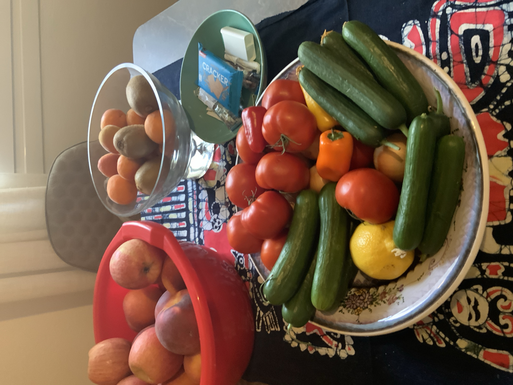 A collection of fruits and vegetables is displayed on a table. In a red bowl on the left, there are peaches and possibly apples. A glass bowl in the center contains apricots and kiwis. To the right of the glass bowl is a green bowl with a blue packet labeled "CRACKER" and a white rectangular object, possibly soap or a small package. In the foreground, on a platter with a patterned cloth underneath, are several cucumbers, tomatoes, a lemon, and a small orange pepper. The setting appears to be indoors, possibly a kitchen or dining area, with a blurred background suggesting furniture and a window. There are no people visible in the image, and no information is available about the specific location within the city of Agios Nikolaos Municipal Unit, Greece, as it is not depicted in the image.