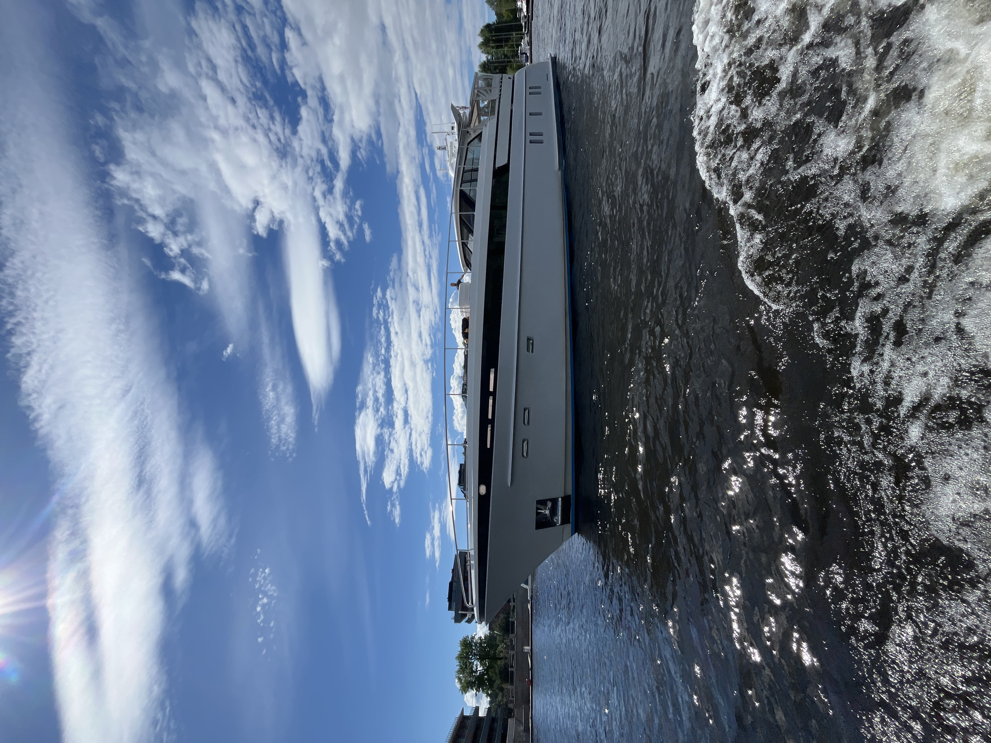 A large white yacht with black accents is cruising on dark water under a bright blue sky filled with white, wispy clouds. The sun is shining brightly, casting a lens flare in the upper left corner of the image. The yacht is the primary subject, positioned in the center of the frame, with its bow on the left and its stern extending to the right. The water around the yacht is choppy, with white foam trailing behind it, indicating movement.
In the background, on the left side of the image, is a modern concrete building with several floors and balconies, possibly residential or commercial. Adjacent to the building are some green trees. Further to the right in the background, more buildings and several other boats are visible along the waterfront. The overall scene suggests a sunny day in an urban waterfront area, likely a canal or river within the city of Saint Petersburg, Russia, given the architectural style of the buildings and the presence of a large yacht. The image captures a moment of leisure or transit on the water.