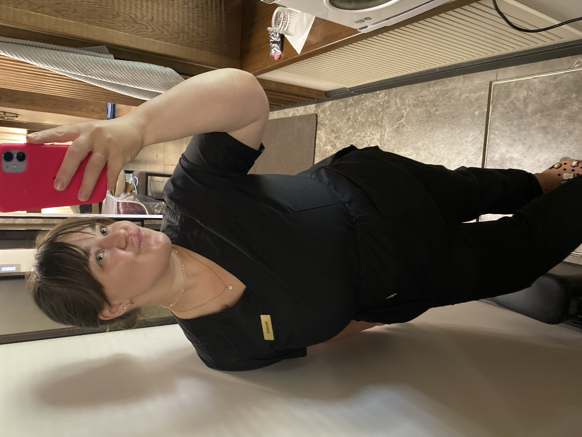 A woman with brown hair, wearing a black scrub top and pants, is taking a selfie in a room. She has a name tag that reads "ANNA" on her left chest. She is wearing a delicate necklace and small earrings. Her left arm is bent, holding a bright pink smartphone. In the background, there is a white wall on the left, and a wooden paneled wall on the right with a towel hanging on it. The floor appears to be tiled. A small table with some items, including a glass and a small package, is visible on the right. The woman is standing in a narrow hallway-like space. There is no indication that this image was taken in the city of Votkinsk, Russia.