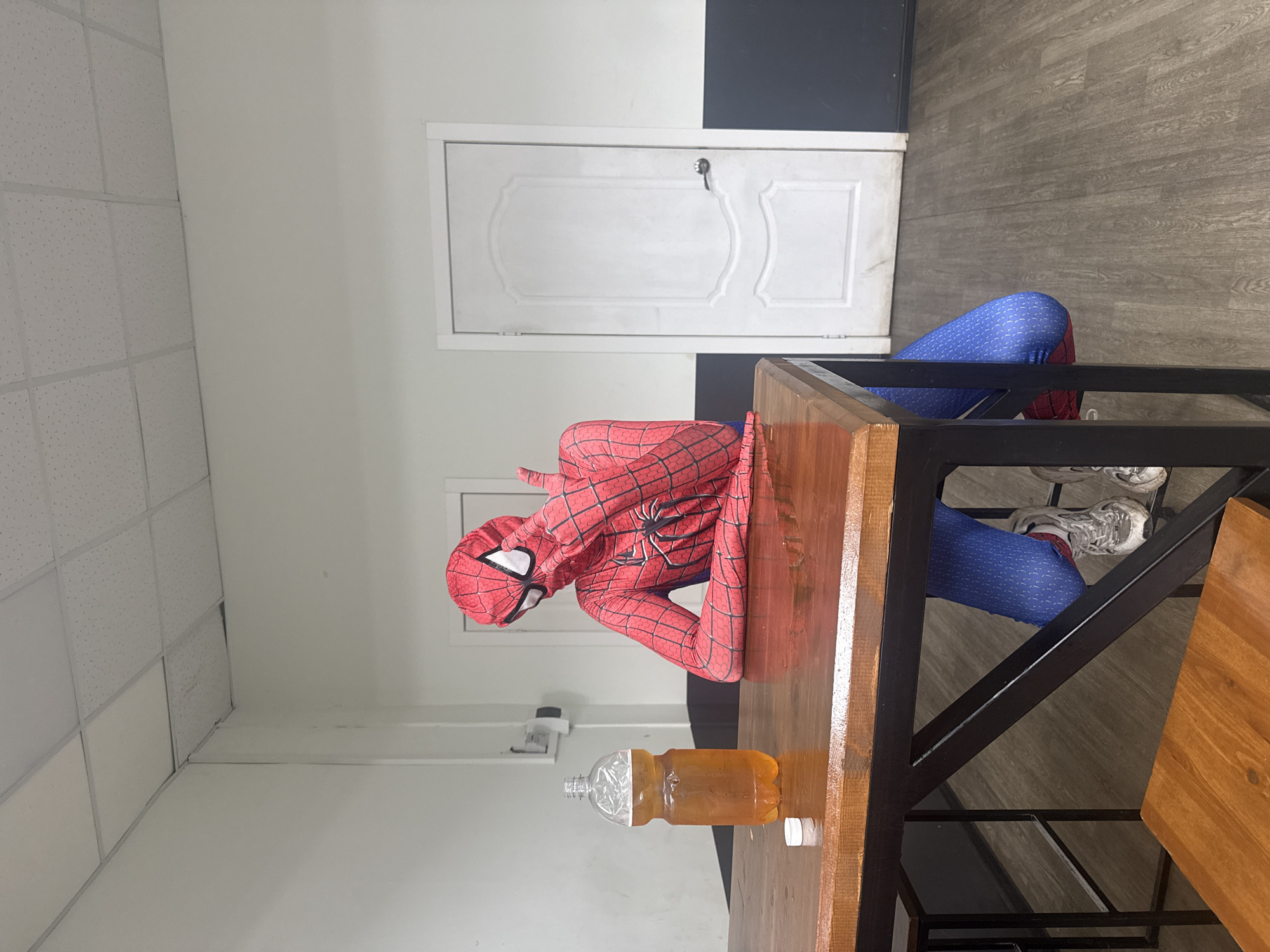 A person dressed in a Spider-Man costume is leaning over a wooden table. There is a large bottle of an amber-colored liquid on the table. The person is wearing gray sneakers. The room has white walls and a drop ceiling. There is a white door with a silver handle in the background. The floor is covered with gray planks. It's unknown if this photo was taken in Votkinsk, Russia.