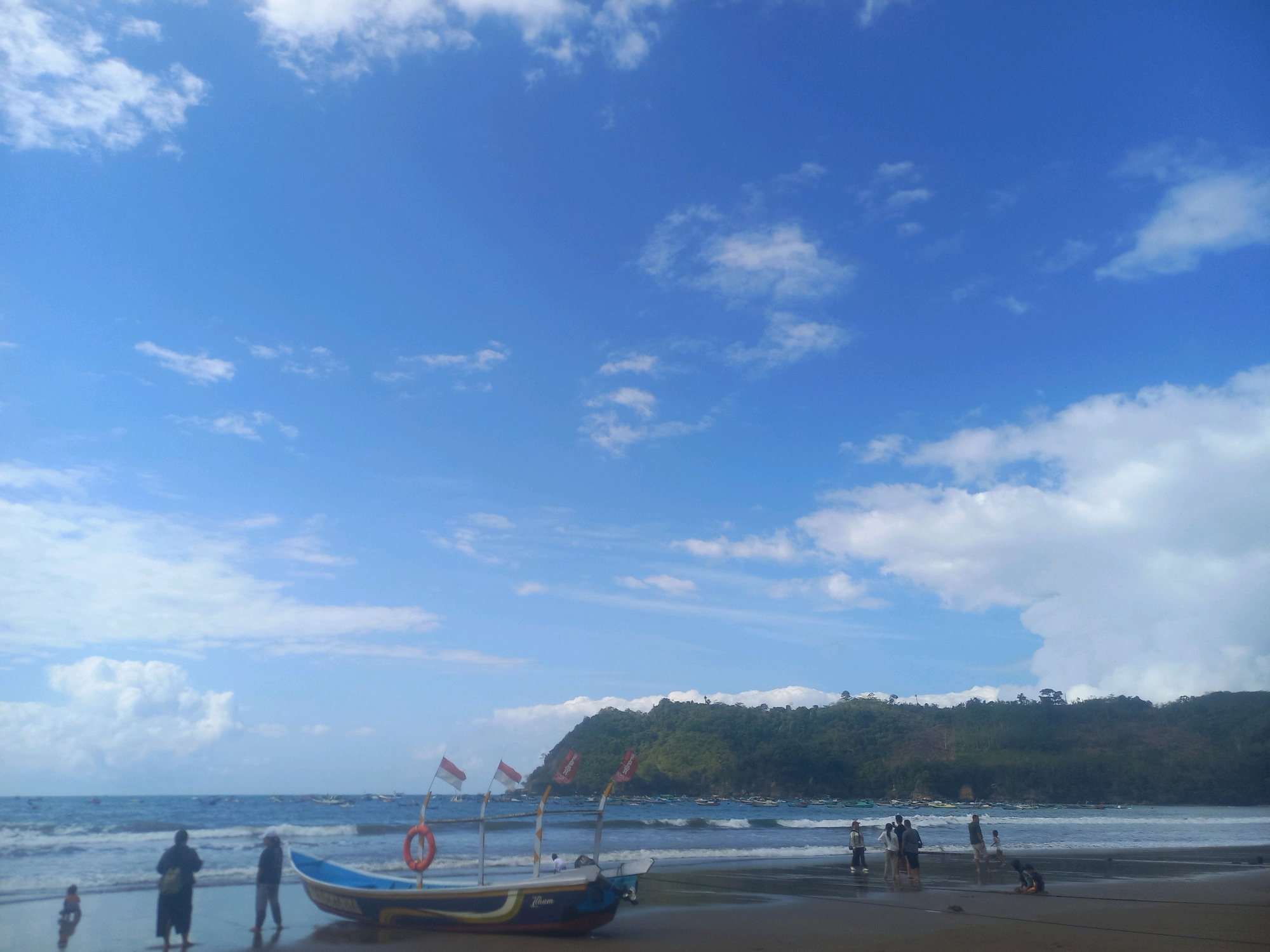This is a wide-angle, eye-level shot of a beach scene in Indonesia. The sky is a vibrant blue with scattered white clouds. In the foreground, the wet sand of the beach reflects the sky. A blue and yellow fishing boat with three Indonesian flags and an orange life preserver is pulled up onto the shore, facing right. Several people are visible on the beach. To the left of the boat, two adults stand facing away from the camera, and a small child is to their left. Further to the right, several more people are standing on the shore. In the middle ground, the ocean has moderate waves, and numerous small fishing boats are scattered across the water. In the background, a lush green, tree-covered hill rises from the coastline. The overall scene depicts a calm, sunny day at a coastal Indonesian village.