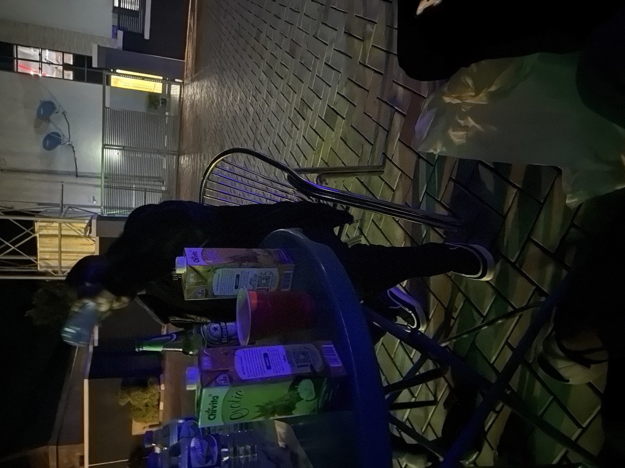 The image depicts a nighttime scene on a paved outdoor patio, likely in Bukuru, Nigeria, as suggested by the context. A person, dressed in dark clothing, is partially visible in the foreground, seated at a blue round table. On the table, there are several items, including two cartons of Chivita juice, a bottle of Heineken beer, a red plastic cup containing a red liquid, and two plastic water bottles. The person is holding a clear plastic water bottle. The background shows a building with a metal gate and illuminated windows, suggesting it might be a residential or commercial property. The lighting is dim, with artificial lights casting colored reflections on the wet-looking paving stones. The overall atmosphere appears casual, possibly indicating a social gathering.