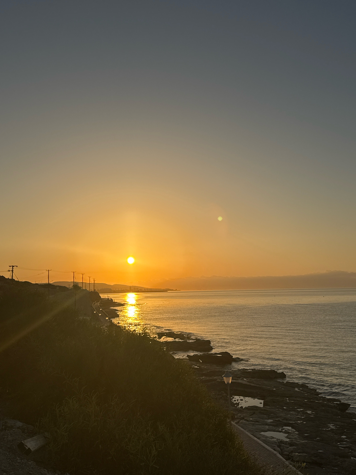 The image depicts a sunrise over the Mediterranean Sea in Agios Tychona Community, Cyprus. The sun, a bright orange orb, is low on the horizon, casting a golden glow across the water and the sky. The sea is calm, with gentle ripples reflecting the sunlight. Rocky shores line the coastline, interspersed with patches of green vegetation. A pathway runs along the coast, illuminated by lampposts. In the distance, a town or city is visible, its buildings silhouetted against the rising sun. The overall atmosphere is serene and tranquil, characteristic of a peaceful dawn by the sea.