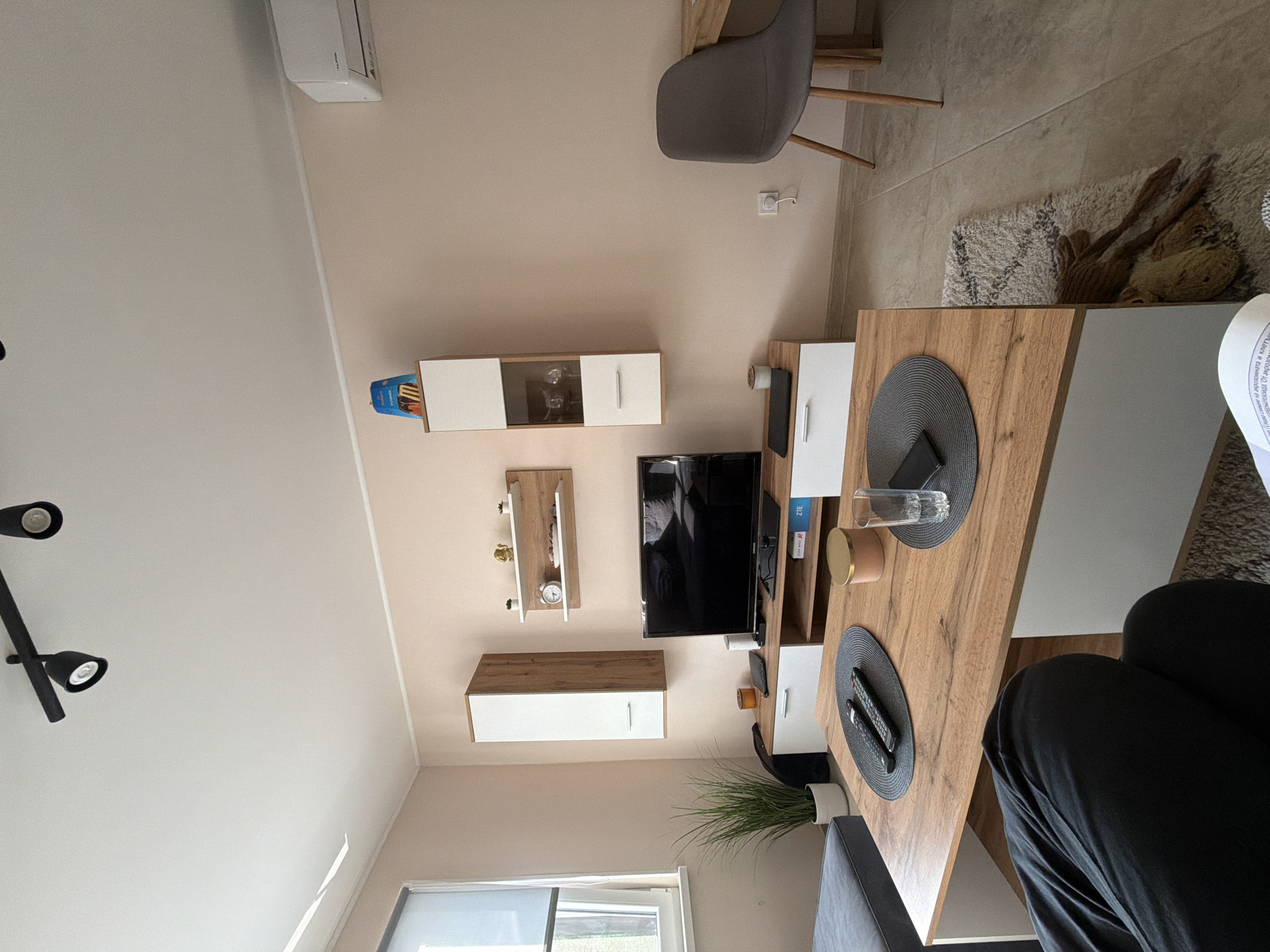 The image shows an interior living room in Litakovo, Bulgaria. The room has a light beige wall with mounted furniture including a white and light wood cabinet, a small shelf with decorative objects, and a larger white and light wood cabinet below the shelf. A black television is mounted between the upper shelf and the lower cabinet. Below the television is a long wooden table with a white cabinet base. On the table are two grey placemats, one with a clear glass and a small tan container, and the other with two remote controls. A green potted plant is near the base of the television stand. A grey chair is partially visible in the upper right. A person wearing black clothing is sitting at the table. A rug is on the floor near the table. A window is visible at the bottom center. A black track lighting fixture is mounted on the white ceiling.