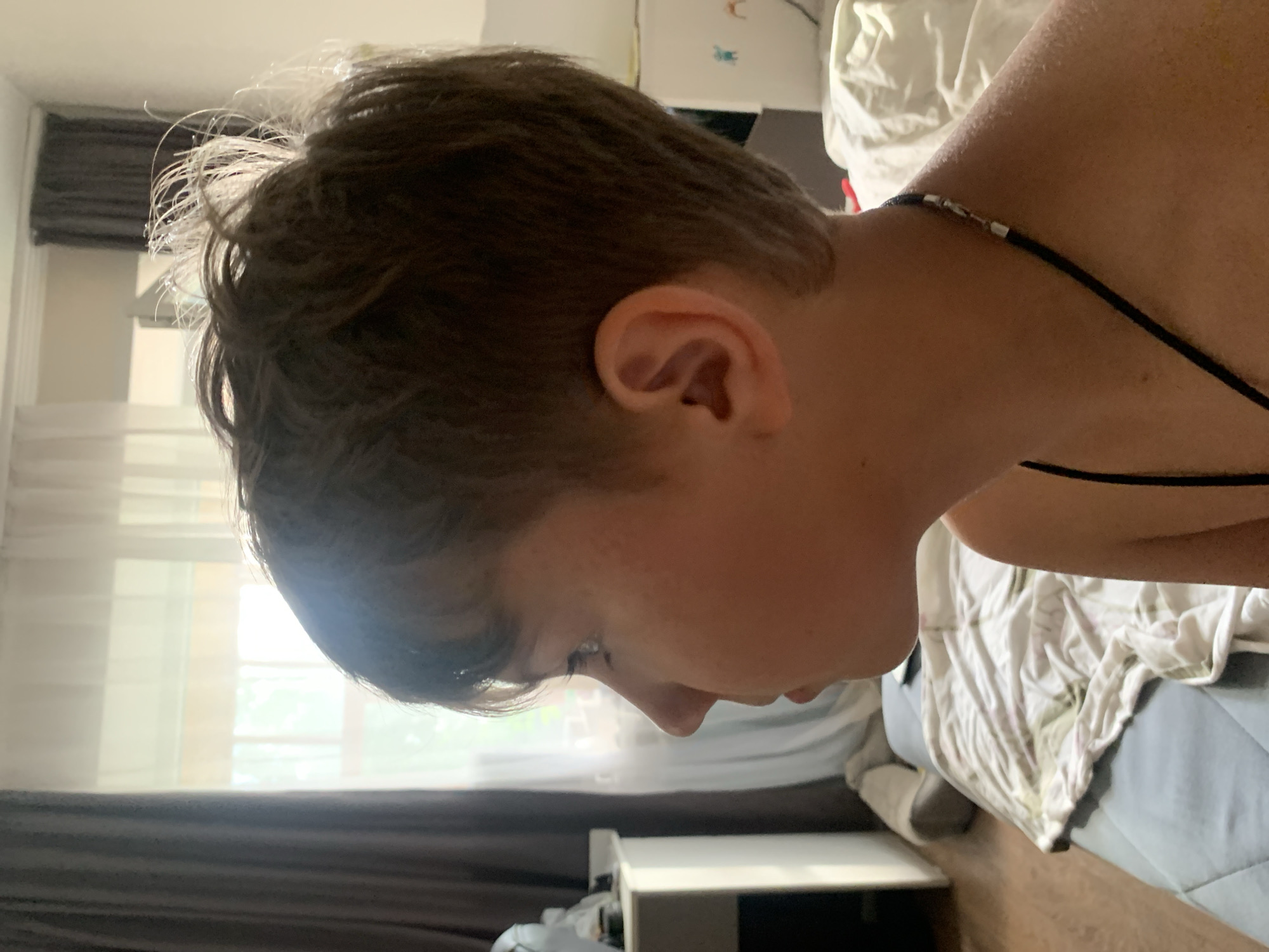 A profile view of a person's head and neck is shown in a close-up shot. The person has short, messy, dark brown hair that appears damp and is styled upwards, with some strands catching the light. The person's face is turned downwards, with their profile visible. The ear is prominent, with its details clearly discernible. A thin black cord with silver clasps is visible around the person's neck and extends down their shoulder. The background suggests an indoor setting, possibly a bedroom. White bedding with some pattern is visible, as is a blue duvet. A dark brown curtain is in the left portion of the frame, and beyond it, a window with sheer white curtains is seen, hinting at natural light filtering into the room. The lighting suggests it could be daytime, possibly morning or afternoon, with soft light illuminating the scene. The image has an informal, candid feel. There is no visible text in the image.