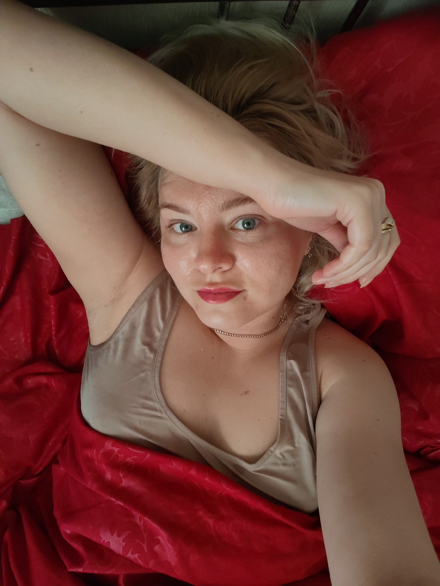 A young Caucasian woman with blonde hair is lying on a bed with red bedding. She is wearing a beige camisole. Her right arm is raised and resting on her forehead, obscuring part of her face. Her left arm is extended down her side, partially covered by the red sheets. She is looking directly at the camera with blue eyes. She is wearing makeup, including lipstick. A gold ring with a stone is visible on her right ring finger. A thin gold necklace is also visible. The lighting suggests it may be daytime, possibly indoors. The overall impression is one of relaxation or perhaps waking up. There is no visible text in the image. The setting appears to be a bedroom in Izhevsk, Russia.