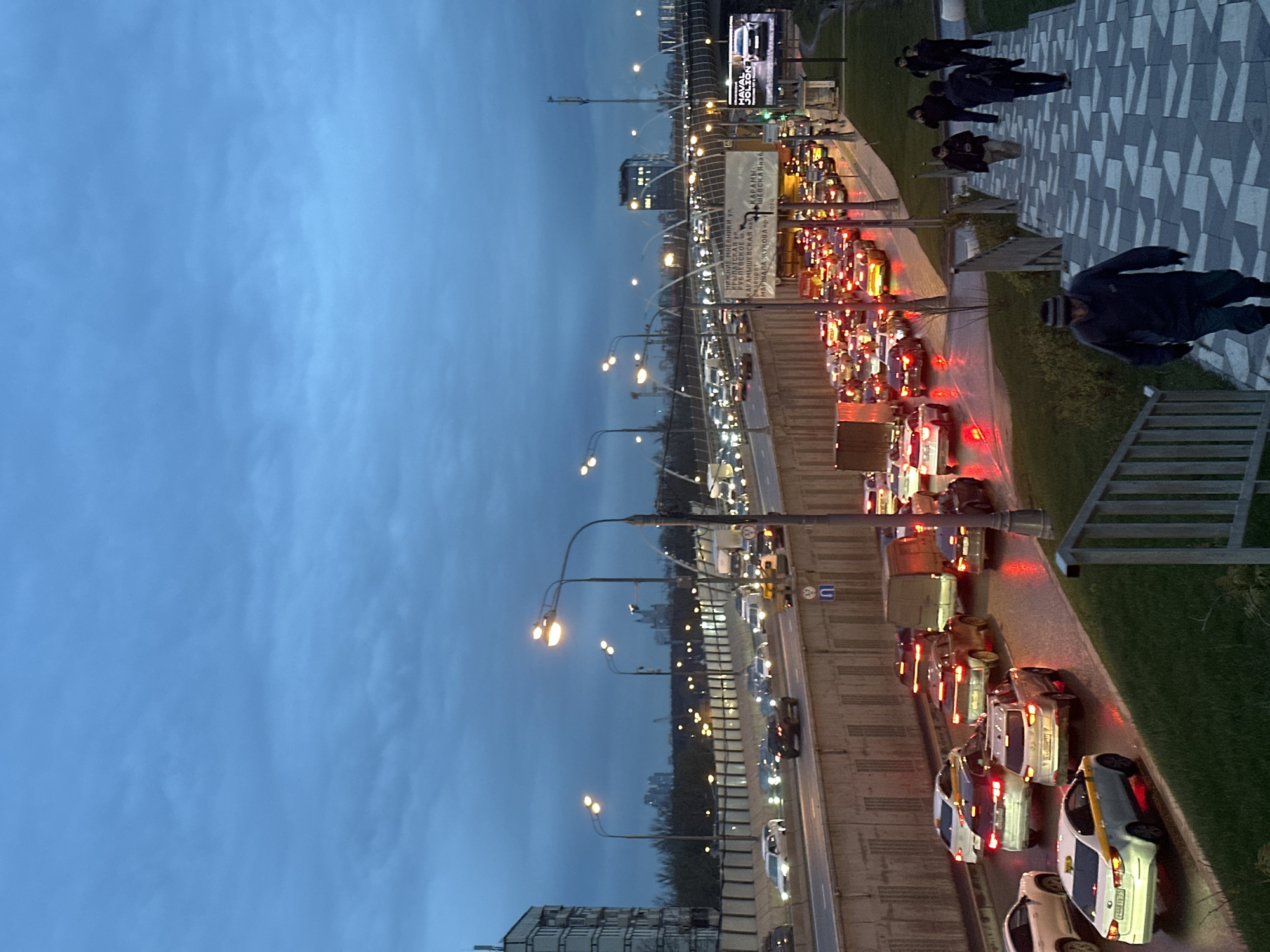 The image shows a highway in Moscow, Russia, in the evening. Traffic is heavy on the road, with many cars queued. Street lights illuminate the scene. Several people are walking on the sidewalk to the right of the highway. A billboard is visible in the distance. The sky is overcast. The photograph appears to have been taken from an elevated position. Signage indicates "МАРШАЛА ЖУКОВА пр-" (Marshal Zhukov Avenue).