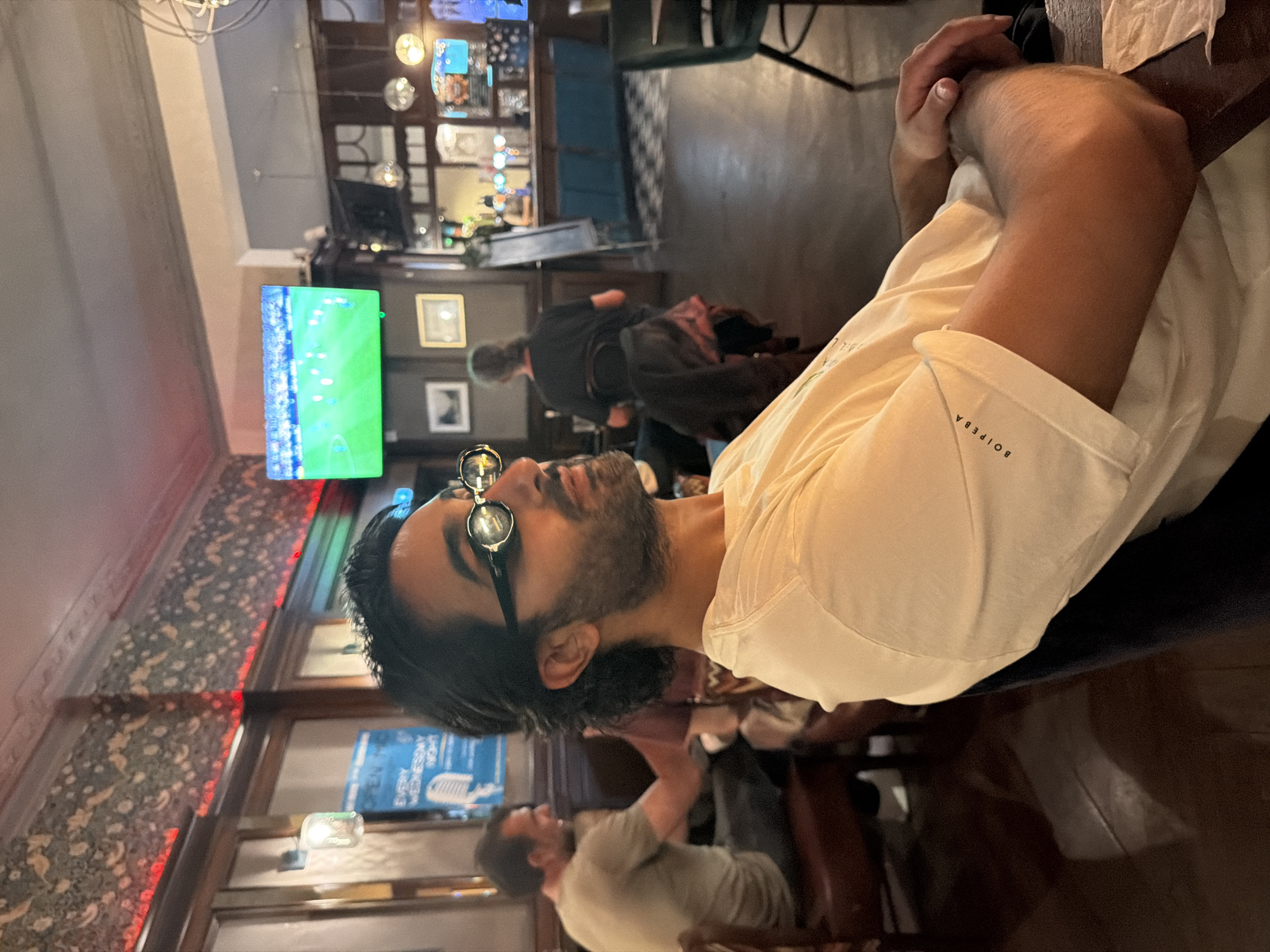 A man with dark hair and a beard is wearing round, steampunk-style glasses and a light-colored t-shirt with the text "BOIPEBA" on the sleeve. He is seated in what appears to be a pub or bar, with wooden furniture and patterned wallpaper visible. In the background, a television screen displays a soccer match, and other patrons are seated at tables and at the bar. The environment suggests an indoor setting with ambient lighting. The man's gaze is directed upwards and to the side, away from the camera.
