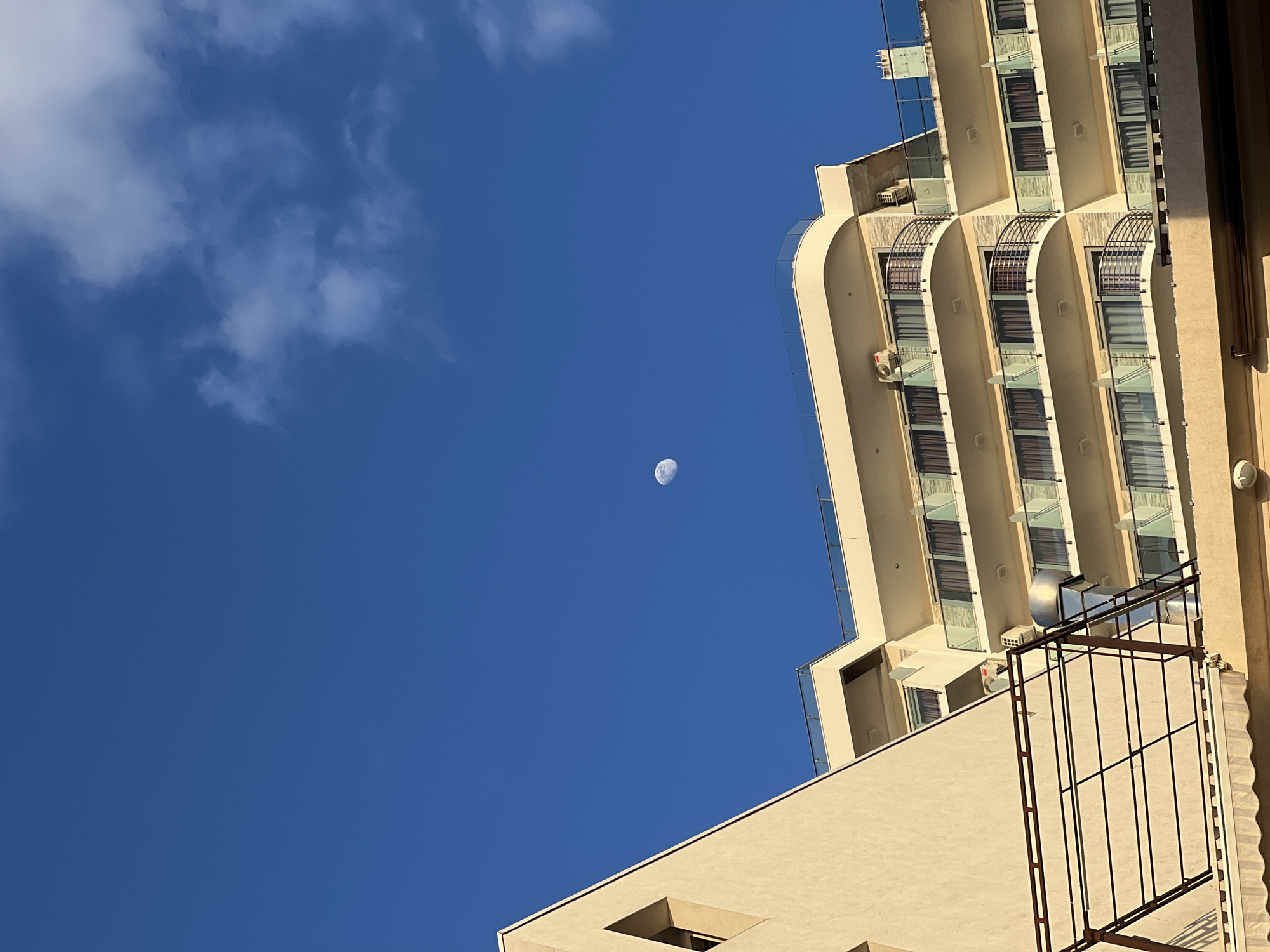 A low-angle shot reveals a cloud-dotted blue sky with a waning crescent moon. The scene also captures the architecture of Budva, Montenegro. To the right, a high-rise building with curved balconies and tinted windows is visible, while a lower beige structure with an open window and a metal grid is in the foreground. The viewpoint is from a position looking upward.