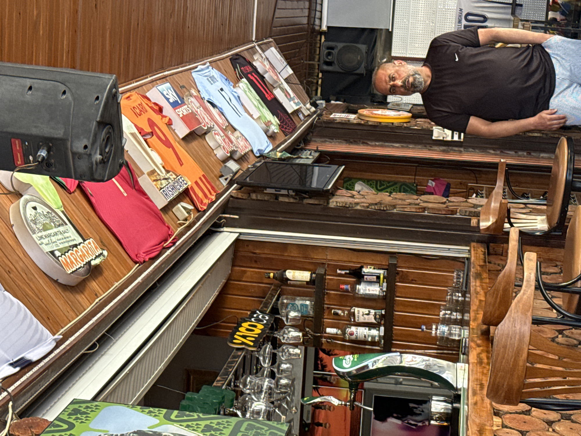 The image depicts the interior of an establishment resembling a sports bar or cafe, possibly located in an unspecified city. A man with a gray beard and wearing a black Nike t-shirt and striped shorts is lying on the bar counter, facing towards the camera. Above the counter, jerseys of various sports teams are displayed, some with associated signage. A speaker is visible above the counter. Behind the bar, shelves with wine bottles and glassware are installed on the wooden wall. Wooden bar stools are present.