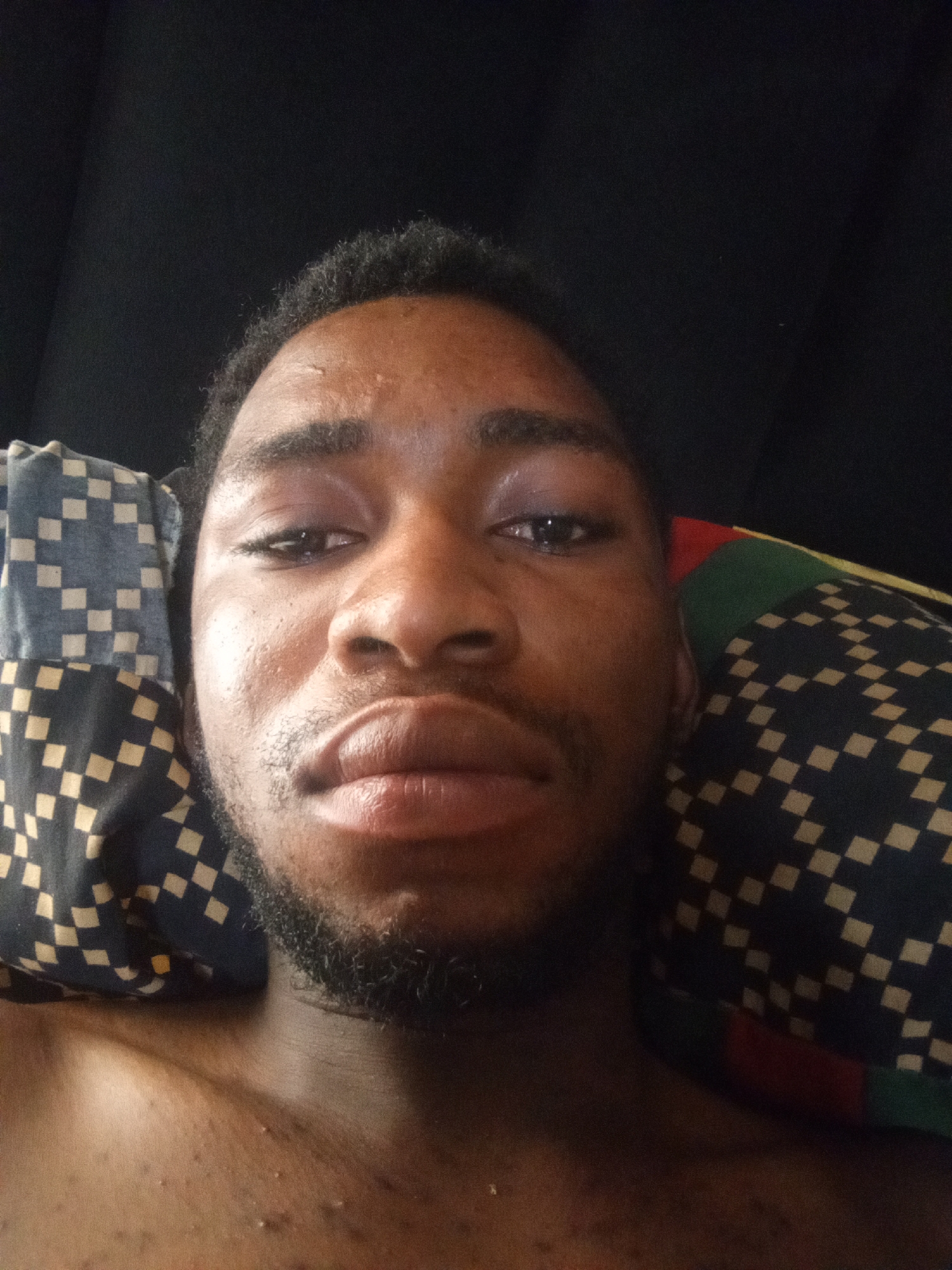 A selfie of a young Black male is taken from a low angle, with his head resting on a pillow. The man has dark, curly hair and dark eyes. His lips are slightly parted, and he has a short beard. He is wearing a dark t-shirt. The background is dark, with a pattern of squares in shades of blue, black, and white on the pillow. The image is dimly lit, with the man's face illuminated by a soft light.