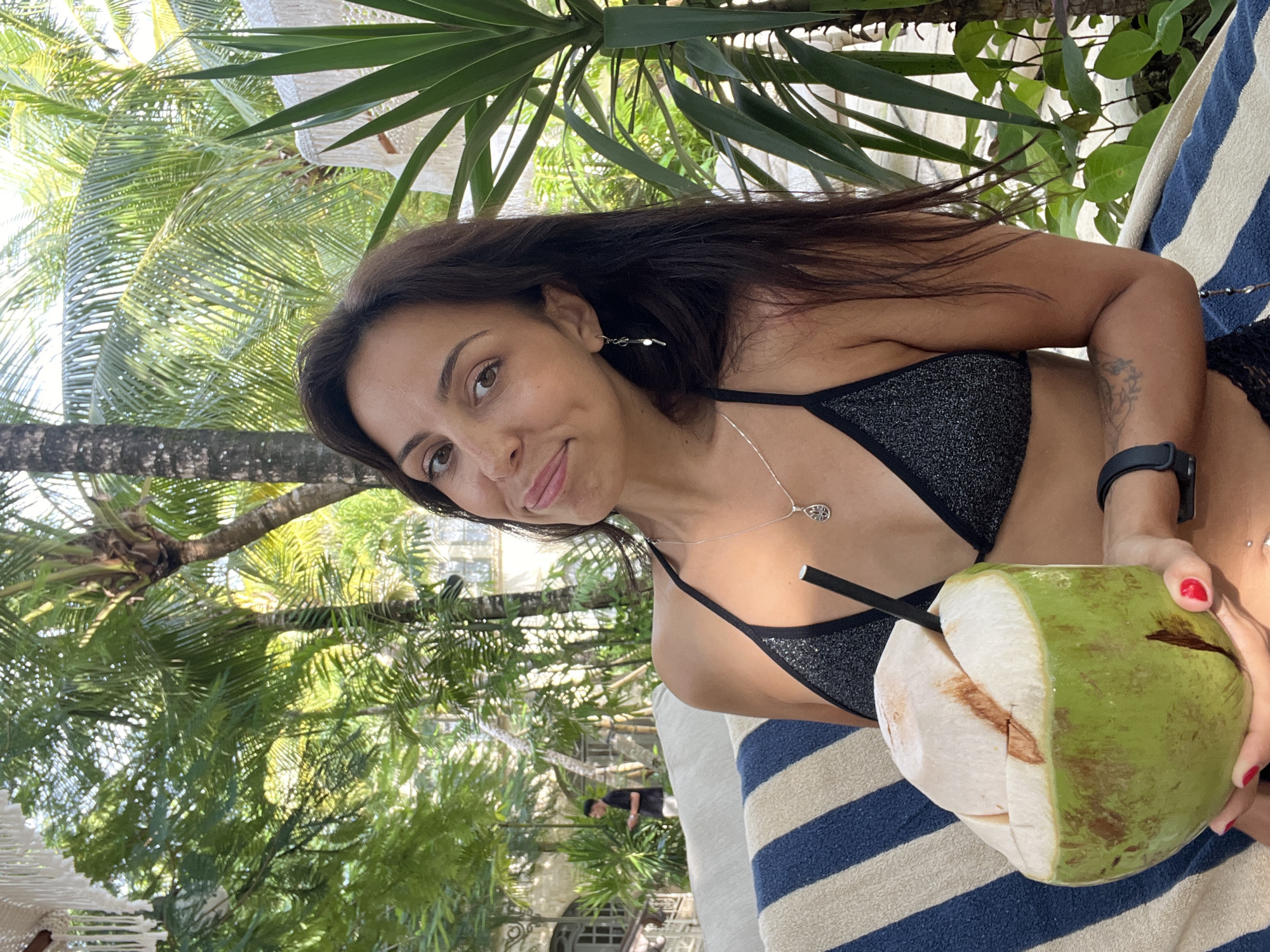 A medium shot captures a woman lounging in a tropical outdoor setting, likely in Kuta Selatan, Indonesia. She has long, dark hair and is looking directly at the camera with a soft smile. She wears a sparkly dark bikini top, a silver necklace with a circular pendant, and matching silver dangle earrings. A black smartwatch is visible on her left wrist, and her fingernails are painted red.
The woman is holding a green young coconut with a black straw inserted into it, suggesting she is enjoying a refreshing drink. She appears to be resting on a striped towel, predominantly blue and white, which is part of a lounge chair or bed.
The background is filled with lush, vibrant green foliage, including prominent palm trees, indicating a sunny, warm, and tropical environment. Hints of white hanging decorations, possibly part of a canopy or outdoor lighting, are visible in the upper left. The overall scene conveys a relaxed and pleasant atmosphere. No visible text is present.