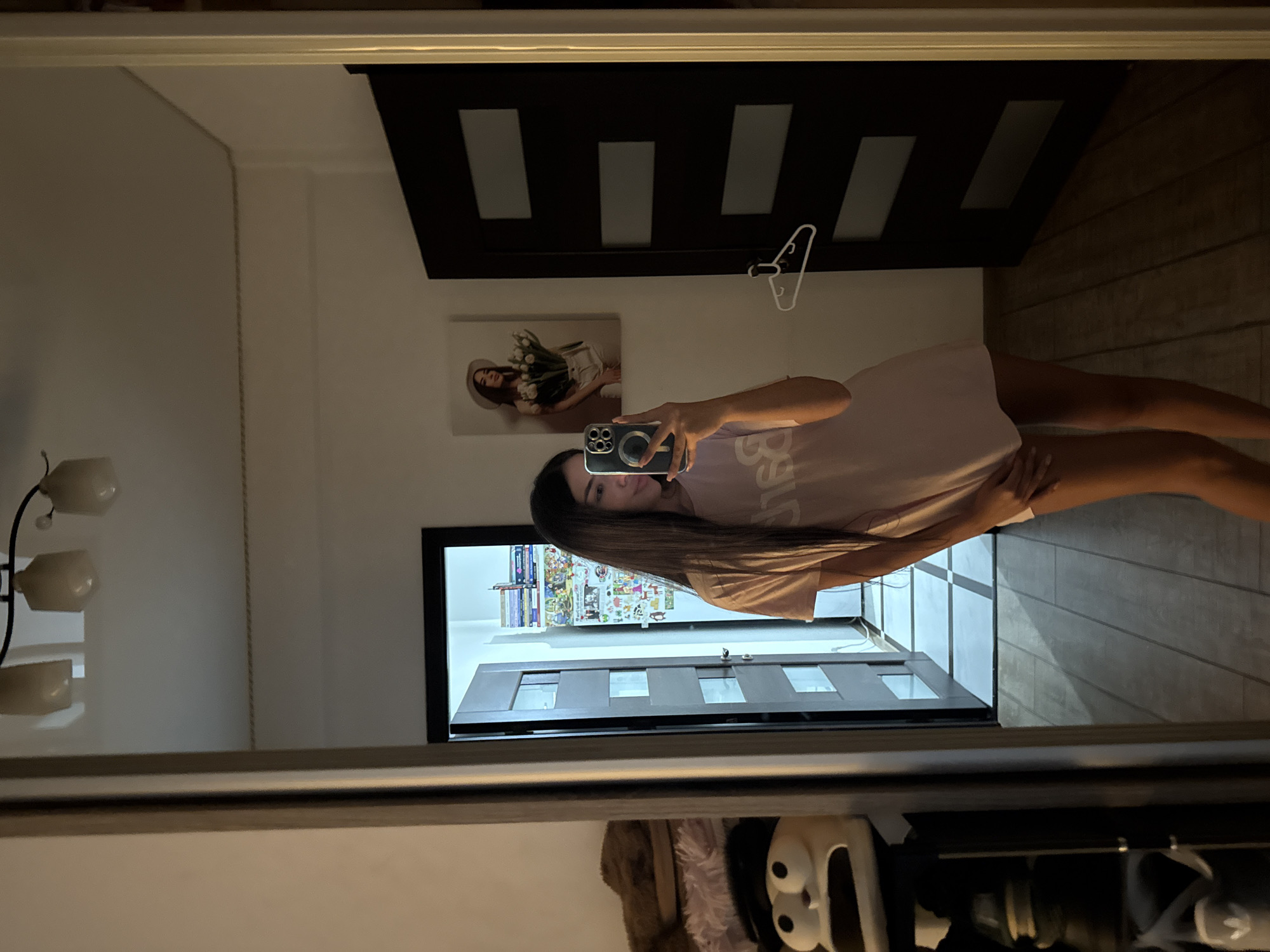 A young woman with long dark hair takes a selfie in a mirror in what appears to be a hallway or entryway of a home. She is wearing a light pink t-shirt that appears to be oversized. The mirror reflects a painting on the wall behind her of a woman holding flowers, and a dark brown door with horizontal rectangular panels. To her left, the mirror also reflects a closet with shelves holding stuffed animals and shoes, and a doorway leading into another room with shelves filled with books and other items. The floor is wooden planks, and the ceiling has lighting fixtures. The overall lighting suggests it is nighttime or a dimly lit room. There is no indication of the specific location within Chișinău, Moldova, beyond it being a residential interior.