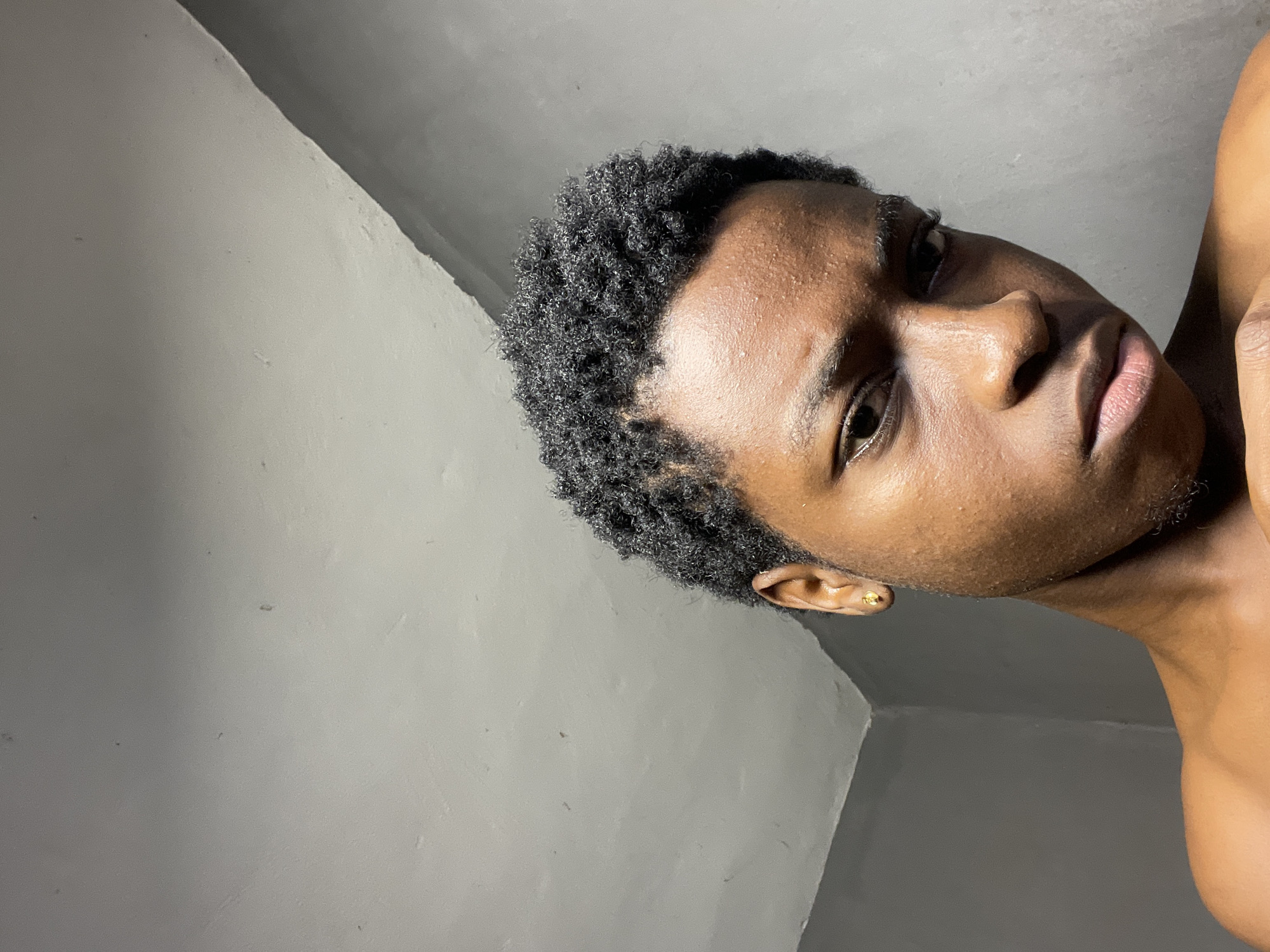 A close-up, slightly canted portrait shows a young Black man indoors in what appears to be Nkpor, Nigeria, as inferred from the user's prompt. He has short, dark, textured hair and is facing the camera with a neutral expression. He is wearing a small, gold-colored earring in his left ear. The background consists of light gray walls meeting at an angle, suggesting a corner of a room. The lighting is somewhat muted, emphasizing the details of his skin and hair texture. The man's shoulders are visible at the bottom of the frame, indicating he is shirtless.