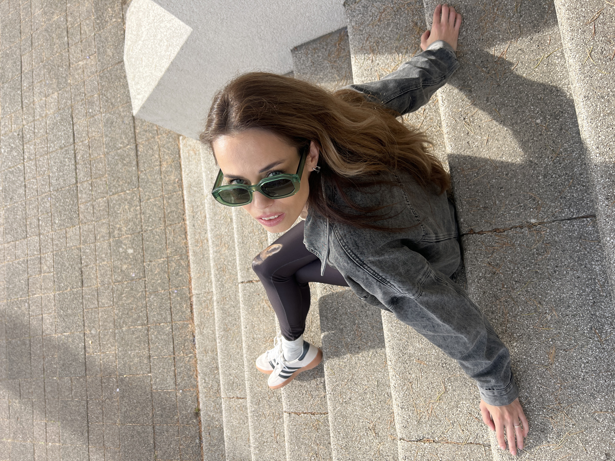 A young woman wearing a gray denim jacket, black leggings, and white Adidas sneakers is sitting on gray concrete steps. She is also wearing green sunglasses. She has shoulder-length brown hair. The steps are part of a larger staircase. The location is outdoor and appears to be in a city, possibly Bar, Montenegro. The ground around the steps is a mix of gray concrete and a cobblestone-like pattern. There are also shadows on the steps.