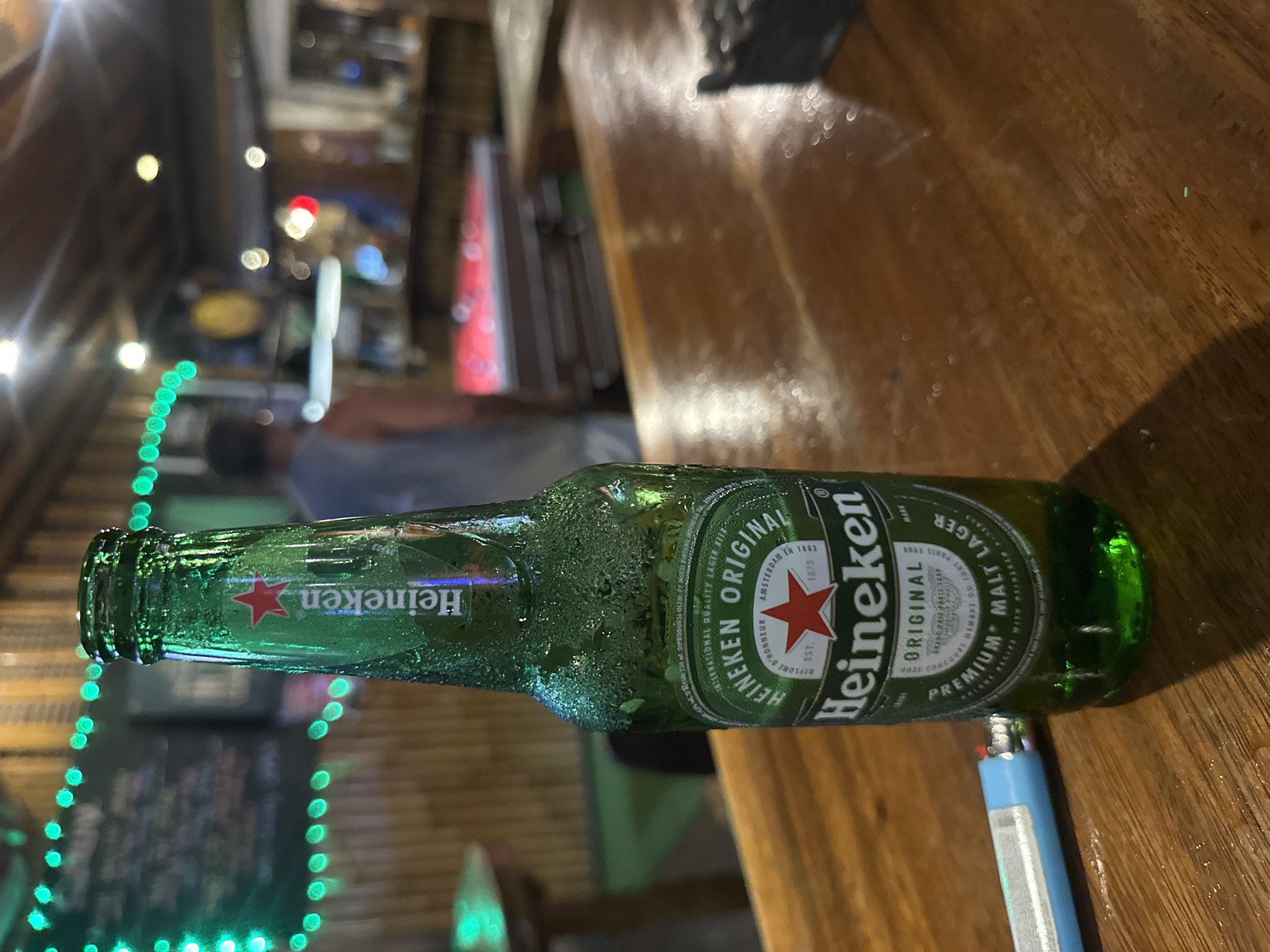 The image depicts a bottle of Heineken beer lying horizontally on a wooden surface, with condensation visible on the bottle. A blue lighter rests near the bottle's bottom. The background shows a blurred scene of what appears to be an outdoor bar or restaurant, with blurry figures of people and strings of green lights. Based on information from the label and potential contextual clues like the presence of a bar setting, the location is inferred to be in North Kuta, Indonesia.