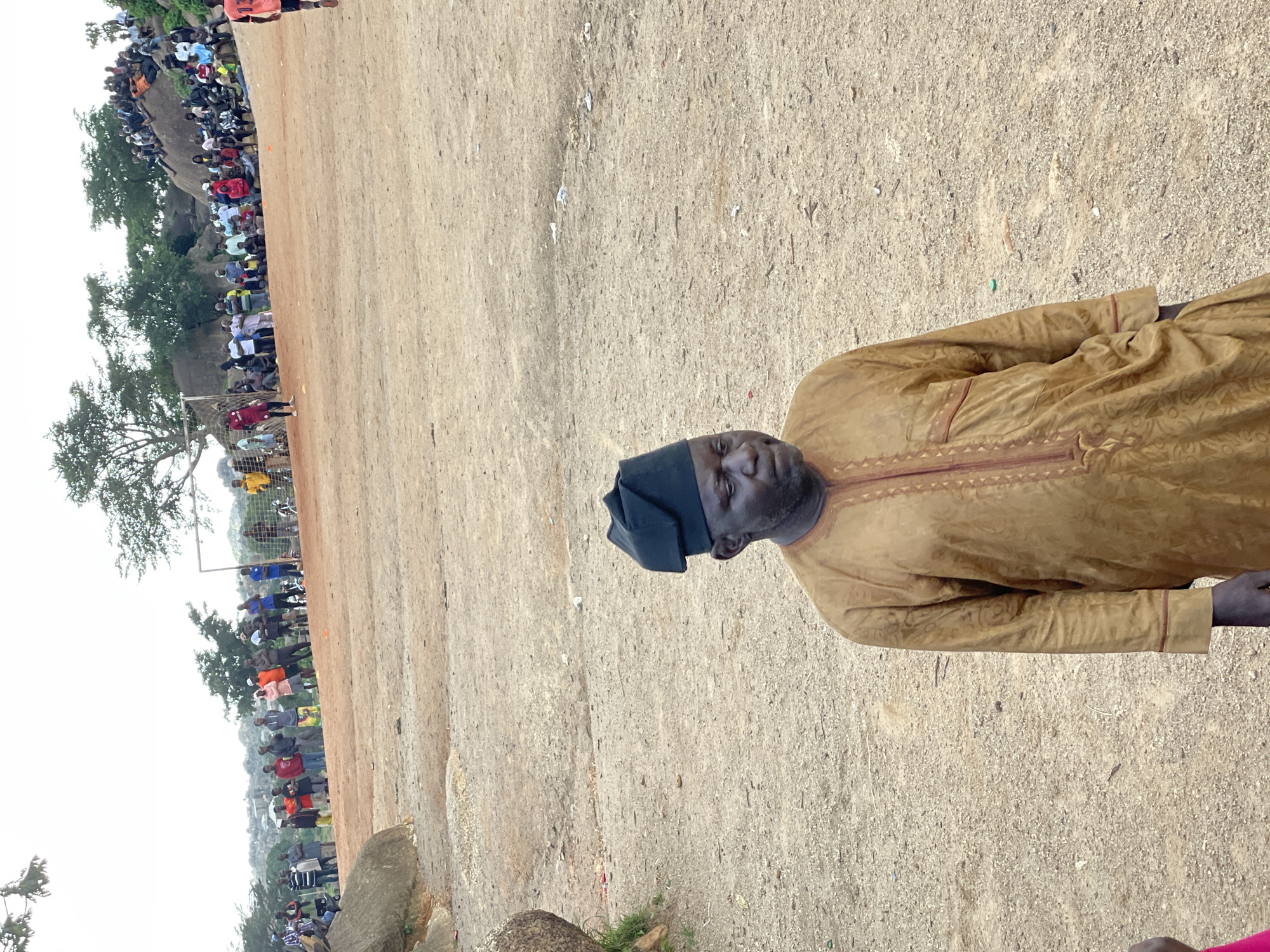 The image depicts a gathering of people outdoors, likely in Jos, Nigeria. In the foreground, a man wearing a traditional tan and maroon patterned robe and a dark cap is the central subject, looking towards the camera. The background shows a large crowd of people of various ages, some standing and others seated, observing an event. Many are dressed casually, with some in bright colors, and a few appear to be wearing football kits.
The scene suggests a public event or gathering, possibly a sporting match or a community festival, given the presence of a football goal visible in the middle ground and the dense crowd. The ground appears to be a dusty, unpaved area, typical of outdoor venues in such regions. Trees and rocky outcrops are visible in the background, providing some natural landscape cues. The lighting indicates it is daytime, with overcast or diffused light, suggesting it could be mid-morning or afternoon. No visible text is present in the image. The overall atmosphere appears to be one of communal engagement and observation.