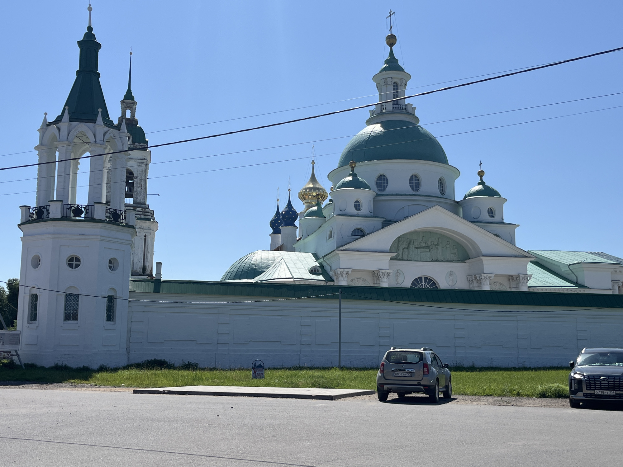 In Kladovitsy, Russia, a sunlit day reveals the Holy Trinity Monastery. The image captures a section of the monastery's white brick wall, topped with a green roof. Adjacent to the wall is a patch of lush green grass. On the right side of the frame, a silver Dacia Duster SUV, license plate beginning with "0838XA", is parked facing left. To its right, a dark grey SUV, possibly a Hyundai Palisade, with license plate beginning with "0718PY", is parked. In the foreground, a paved road stretches across the bottom of the image, with a thin, grey curb separating it from the grassy area. Power lines crisscross the bright blue sky above. The monastery itself features a prominent white bell tower with a dark green spire on the left, and a larger, white building with multiple green domes and golden crosses on the right. The architectural style is distinctly Orthodox Christian.