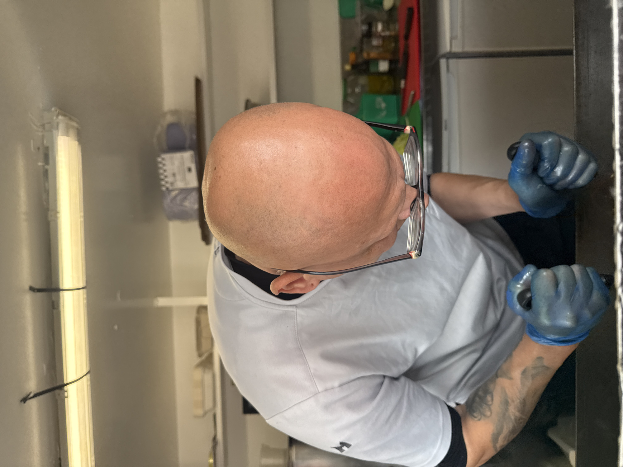 An overhead shot captures a bald man wearing a white chef's jacket and blue gloves, leaning over a counter. His head is bent down, with glasses resting on his nose and the frames hanging low. His left arm, adorned with a tattoo, is visible as he holds something in his gloved hand. The background suggests a commercial kitchen environment with shelves, containers, and bottles. The scene takes place within a food truck or catering unit located in Tewkesbury, United Kingdom, identifiable by the distinctive lighting and the layout of the workspace.