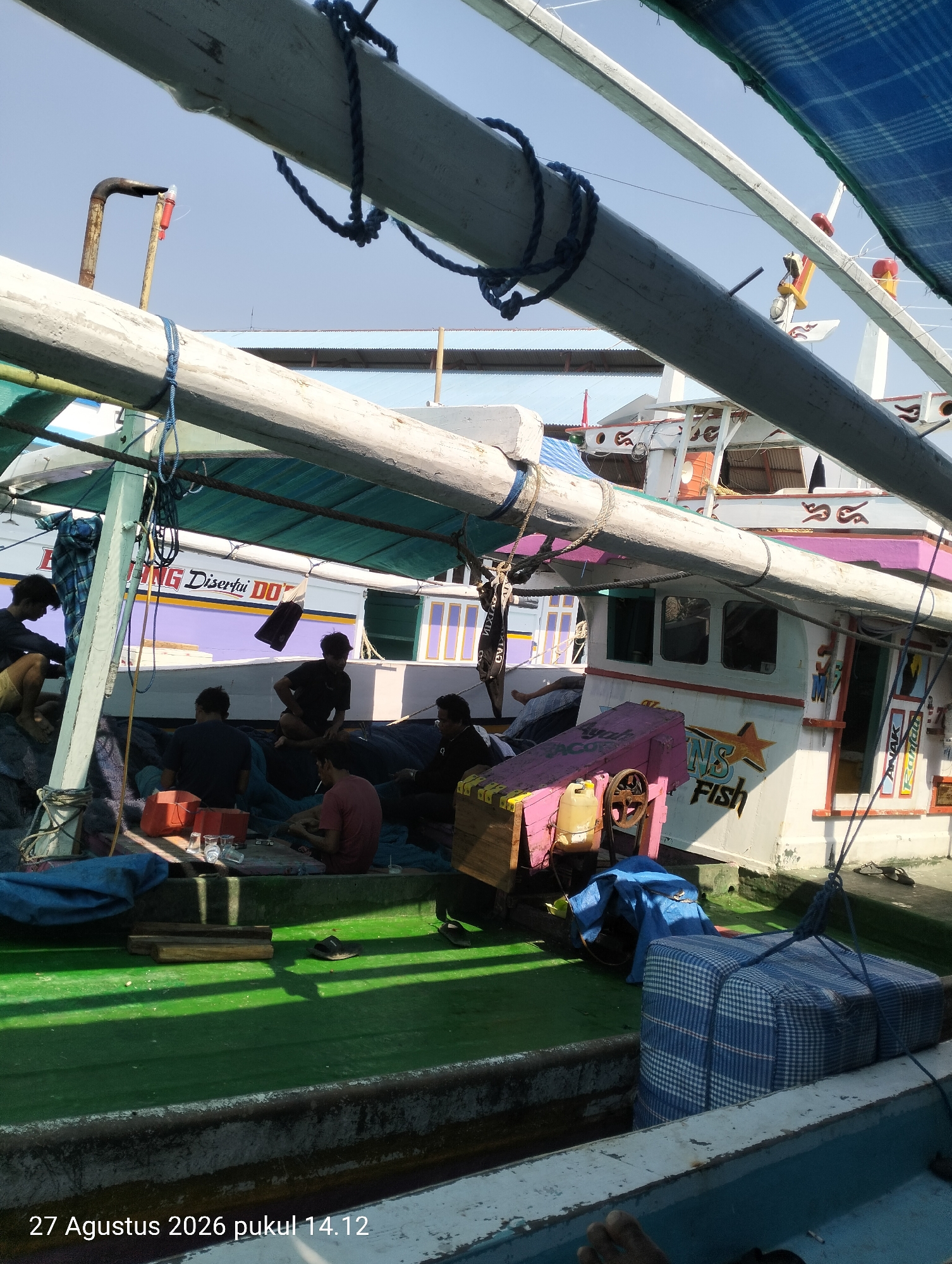 The image captures a scene on a fishing boat in Kendari, Indonesia. Several individuals are gathered on the deck, some sitting and interacting, while others appear to be engaged in work or rest. The boat itself is equipped with fishing gear and appears to be docked or anchored. The environment suggests a sunny day, with clear skies visible. The presence of fishing equipment and the overall setting point towards the likely location being a harbor or a fishing village in Kendari.