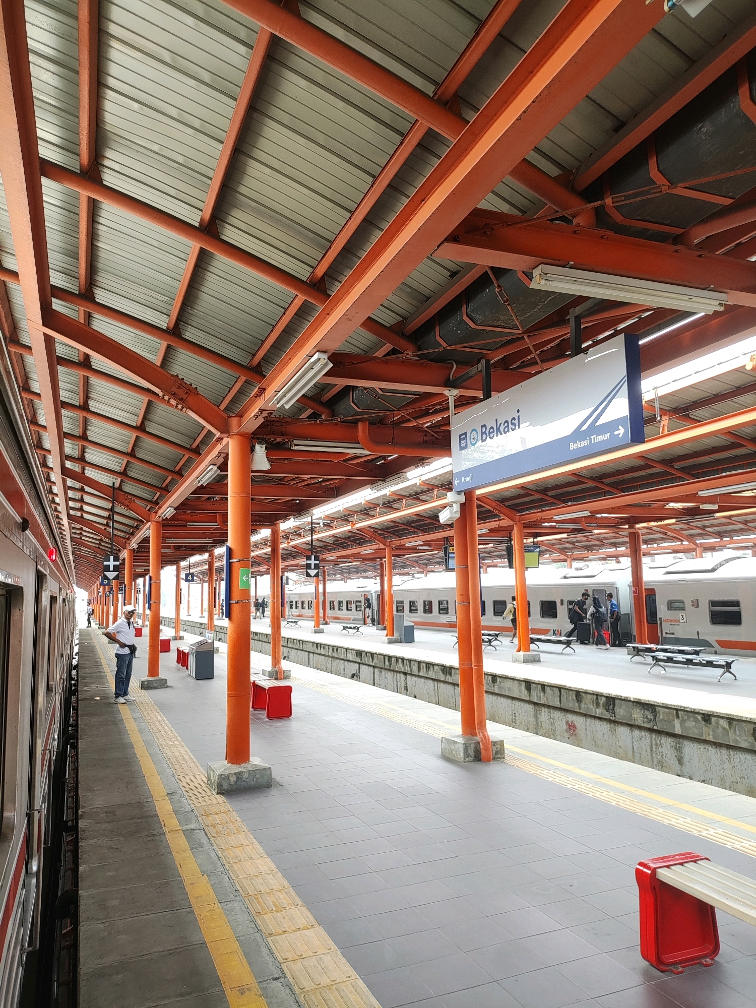A train station platform in Bekasi, Indonesia, is depicted. The platform is covered by a metal roof supported by orange, I-beam-like structures. The roof is corrugated and appears to be made of galvanized steel. Fluorescent lighting fixtures are attached to the underside of the roof. Orange cylindrical pillars support the roof at intervals along the platform.
The platform itself is paved with grey tiles. A yellow tactile paving strip runs along the edge of the platform next to the train tracks. Red, metal benches with slatted seats are positioned on the platform. A grey trash bin is also visible.
A train with a white and red exterior is stopped at the platform. The train appears to be a commuter or intercity train. Several people are visible on the platform. In the foreground, a man in a white shirt and blue jeans stands near the train, holding a small bag. Further down the platform, a group of people are standing near another train.
A blue and white sign hangs from the roof, indicating "Bekasi" and providing directional arrows for "Bekasi Timur" (East Bekasi) and "Kranji." The architecture and signage suggest this is a functional, urban train station. The environment is clean and orderly.