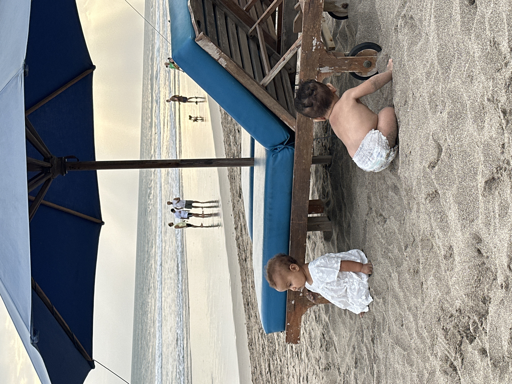 This is a vertical, outdoor, candid photograph taken on a beach in North Kuta, Indonesia. The image captures two young children playing in the sand next to a blue-padded lounge chair. In the foreground, a dark blue beach umbrella is partially visible on the left side. The sun appears to be low in the sky, casting a soft, warm light, suggesting it is either early morning or late afternoon. The sky is bright and overcast.
The child in the foreground, closer to the lounge chair, is wearing a white lace dress and is seen from the side, looking down at the sand. Their hands are resting on the wooden structure of the lounge chair. The second child, further back and to the right, is wearing a diaper and is in a crawling or kneeling position, facing away from the camera. Their back is towards the lounge chair.
In the background, the ocean and the beach are visible. Several people are walking along the shoreline. One group of three individuals is in the middle distance, appearing as silhouettes against the bright water. Another silhouette of a person is further down the beach. The overall atmosphere of the image is peaceful and relaxed, indicative of a family vacation or a leisurely day at the beach. There is no visible text in the image.