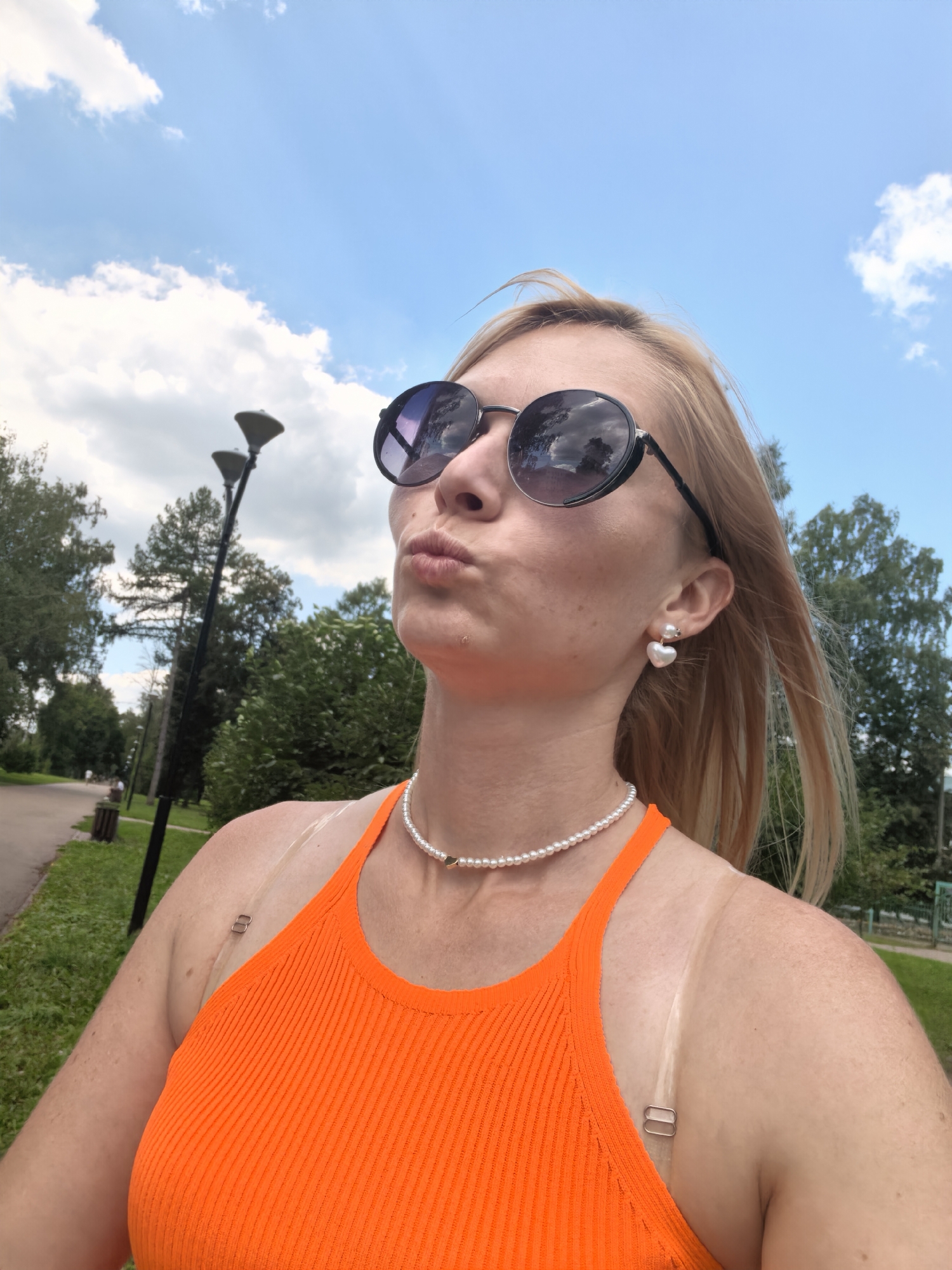 The image shows a woman with blonde hair and sunglasses, wearing an orange tank top and a pearl necklace. She is outdoors, with trees and a blue sky with clouds in the background. There are lampposts visible in the distance. The woman is looking up and to the side, with her lips pursed. There are visible tan lines on her shoulders. The location is identified as Sterlitamak, Russia.