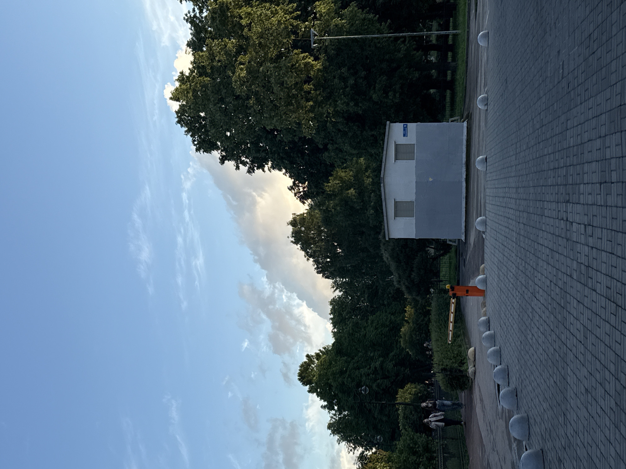 The image depicts a scene in a park-like area, possibly within the city of Saint Petersburg, Russia, given the aesthetic. The sky is a mix of clear blue with wispy white clouds, and some larger, darker clouds, suggesting late afternoon or early evening light. Dominating the right side of the frame are large, dense green trees. In the mid-ground, a small, rectangular building with a gray base and white upper section, featuring two windows, is visible. Adjacent to the building, a modern streetlamp stands tall. In the foreground, a paved area, likely a parking lot or walkway, is marked with a pattern of interlocking gray pavers. Several rounded, gray concrete bollards are lined up along the edge of the paved area, serving as barriers. To the left of the bollards and paved area, a road or path is visible, lined with a dark metal fence. On this path, two individuals, appearing to be young women, are walking away from the camera. One is wearing a light-colored jacket and dark pants, and the other is in a darker jacket and jeans. Further back, an orange automatic barrier arm is extended across the path. The overall atmosphere is tranquil, with a blend of natural elements (trees, sky) and urban infrastructure.