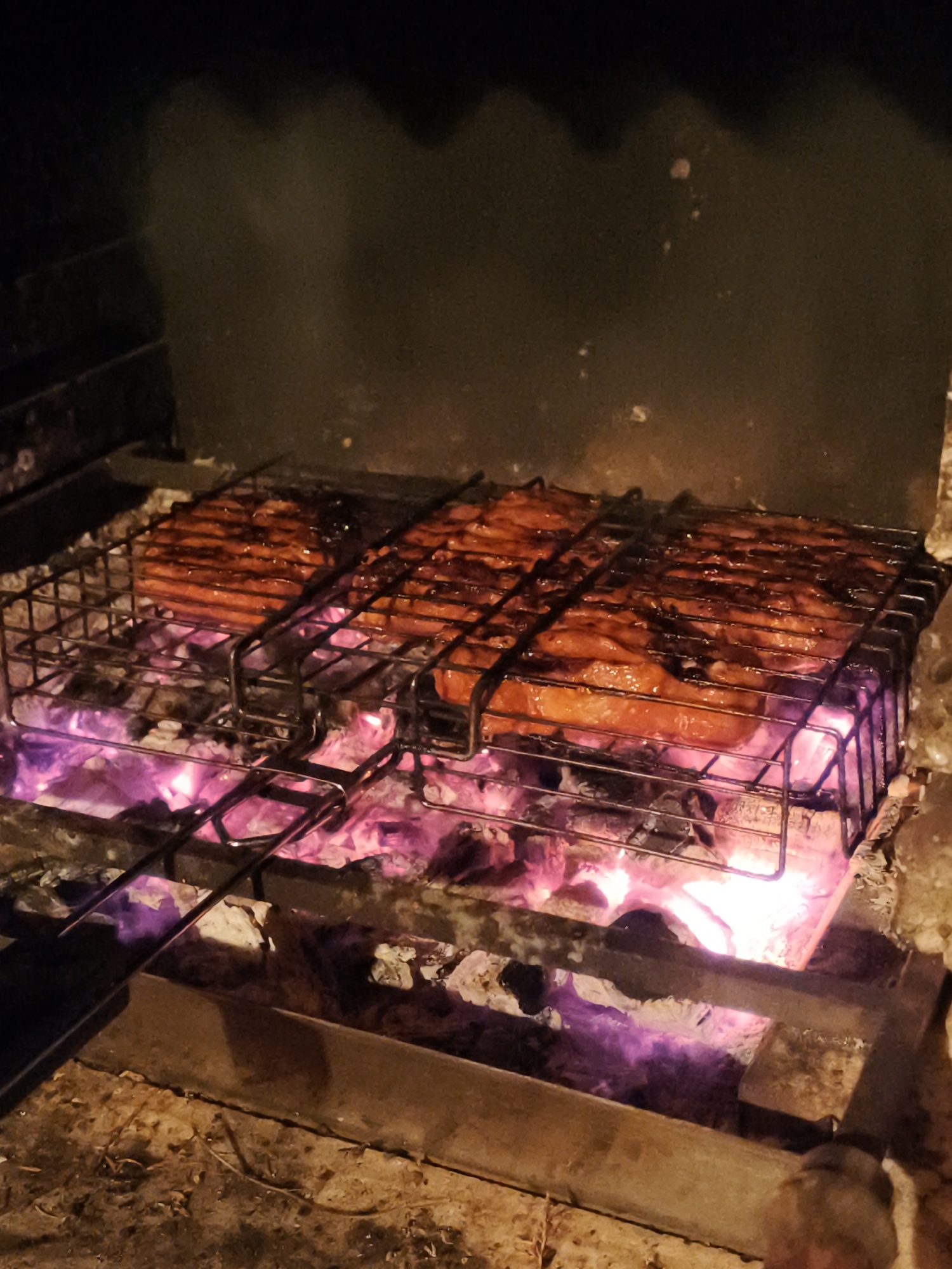 The image depicts a close-up view of three pieces of meat, likely sausages, cooking on a metal grill grate over a bed of glowing pink and orange embers. The meat appears to be glazed and is arranged in a row on the grill. The grill itself is resting on a black metal structure, which is part of a barbecue or fireplace. The background is dark and out of focus, suggesting an outdoor setting at night, possibly in the city of Angarskie Khutora, Russia. There are no people visible in the image. The focus is entirely on the food being cooked over the fire.