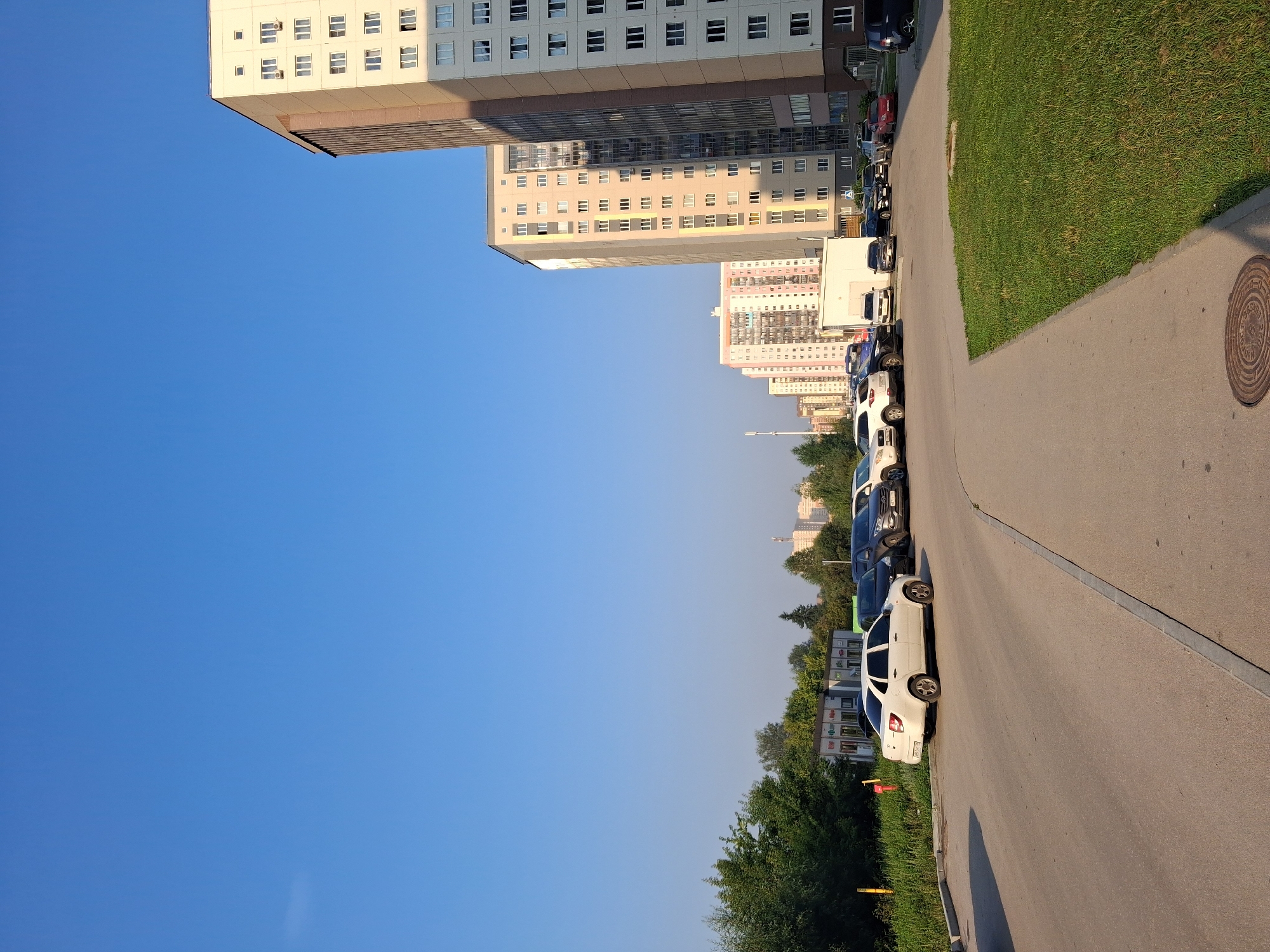 The image captures a wide-angle view of a residential area in Novosibirsk, Russia, on a clear day. The scene is dominated by a bright blue sky and features several multi-story apartment buildings. In the foreground, a paved road curves through a parking lot filled with various cars, including a white sedan, dark SUVs, and a red car. To the left of the road, a strip of green grass and trees borders the area. In the background, more apartment buildings are visible, some with colorful facades. The buildings appear to be typical of Soviet-era or post-Soviet era urban development. The lighting suggests it is either morning or late afternoon, with shadows cast by the buildings and trees. There are no people visible in the frame. A manhole cover is present on the paved area in the lower right portion of the image.