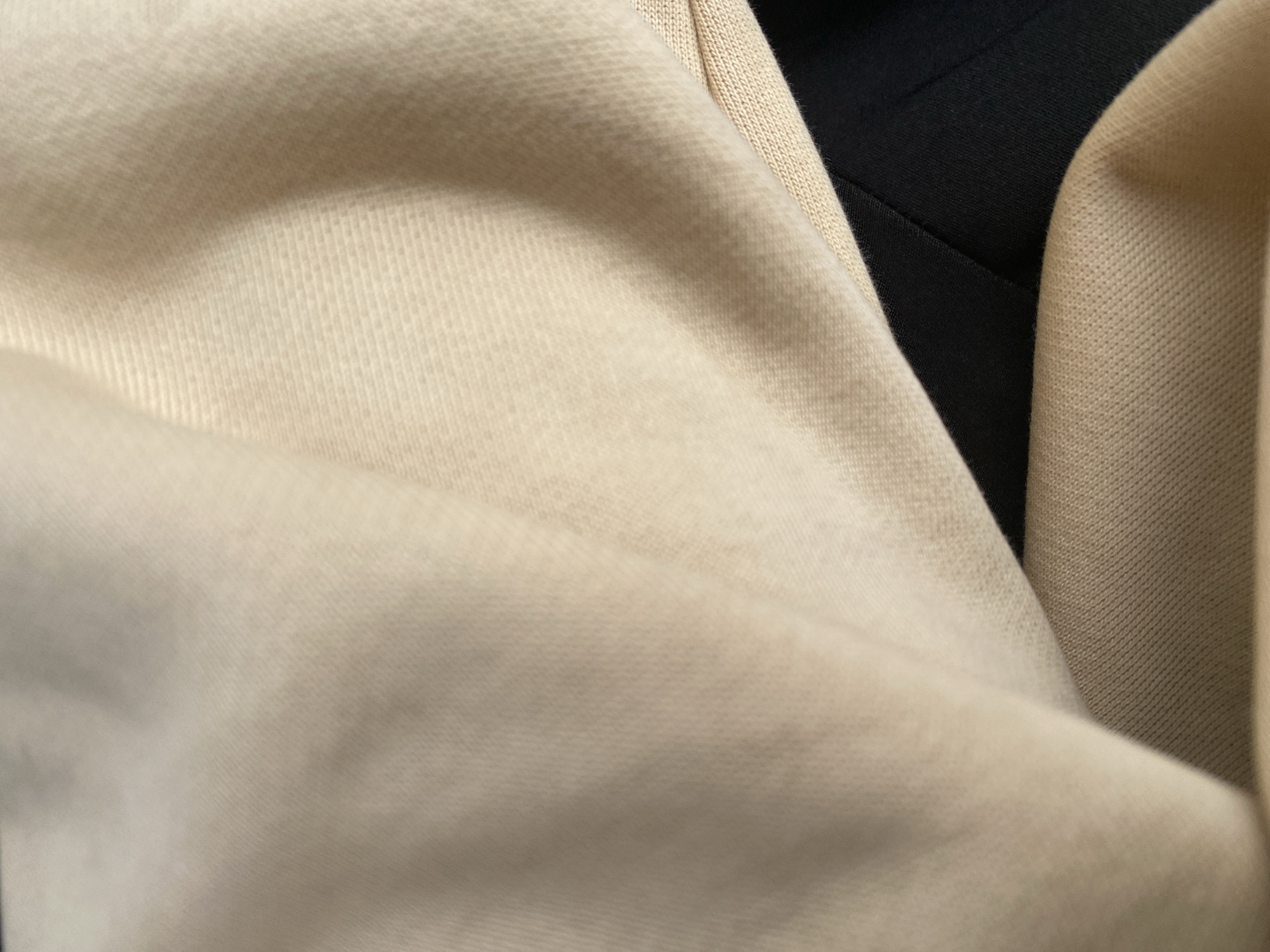 The image is a close-up shot of fabric textures. The primary subject is the texture of a light beige, soft fabric with visible knitted lines. This fabric is folded and draped, creating soft curves and shadows. To the right and in the upper background, a darker fabric, appearing black or very dark navy, is visible, also folded. A narrow strip of a textured beige fabric, possibly a different weave or material, is seen between the lighter beige and the dark fabric in the upper right portion of the image. The lighting is soft and diffused, creating subtle highlights and shadows that emphasize the texture of the materials. There is no discernible activity or event taking place, as the image focuses solely on the textiles. No people, text, time of day, weather, or specific location cues are present. The context provided (Jalingo, Nigeria) does not offer visual information within the image itself.