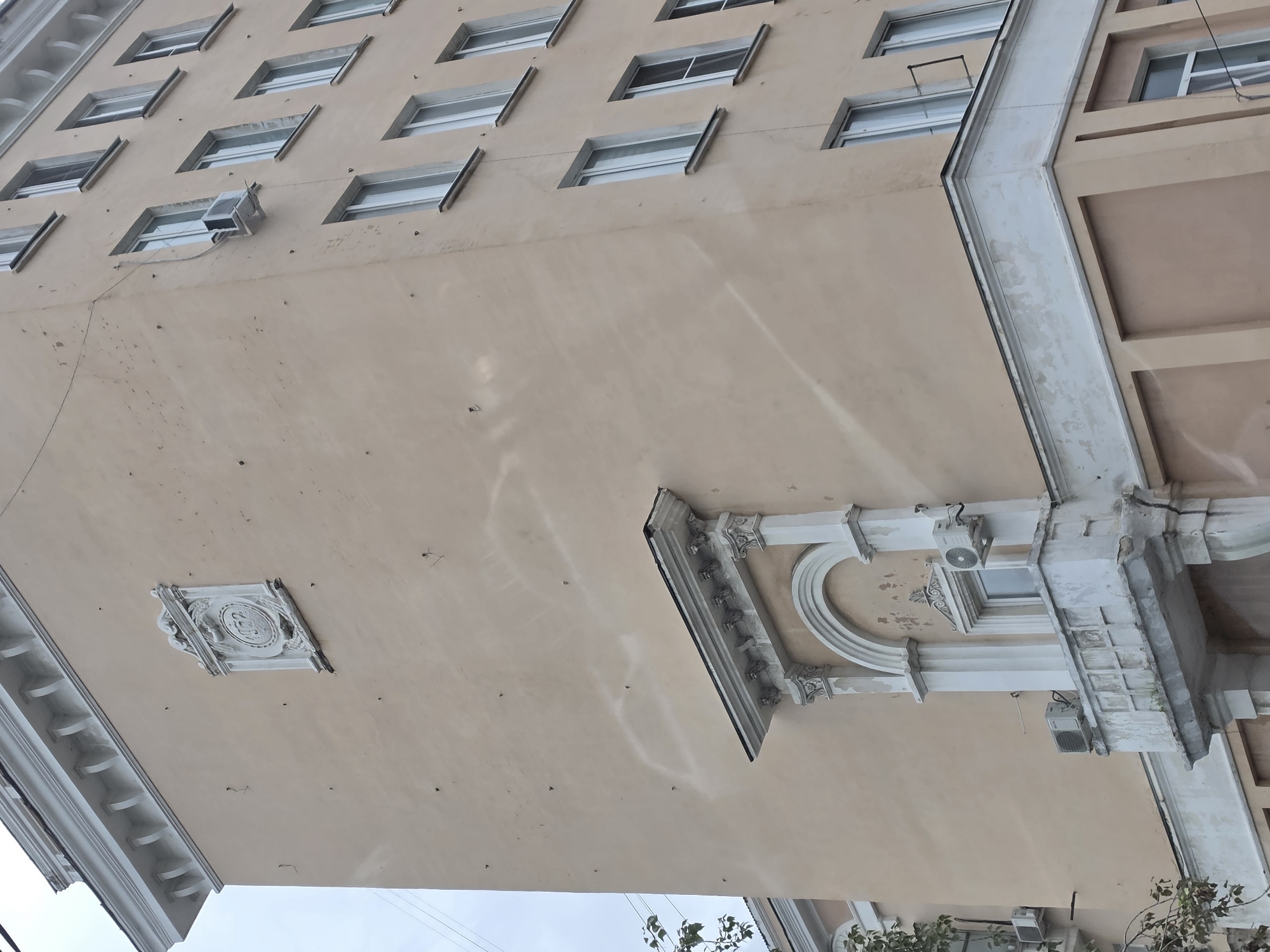 The image shows the facade of a multi-story building in the city of Nizhny Novgorod, Russia. The building is painted a light peachy color and features several architectural elements, including decorative molding, a balcony with ornate railings, and a relief plaque with the year "1952" visible. There are numerous small dark marks on the facade, possibly bullet holes or shrapnel damage, which are particularly concentrated on the upper portion of the building. Several windows are visible, some equipped with air conditioning units. A tree with sparse foliage partially obscures the lower left corner of the building. The sky is overcast.