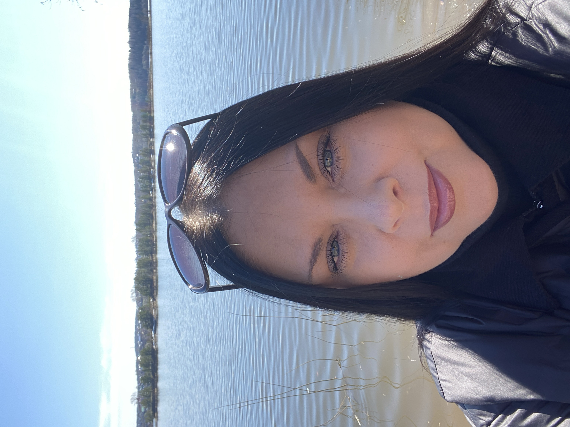 A young woman is posing for a selfie in an outdoor setting. Her sunglasses are perched on top of her head. The background features a body of water, possibly a lake, and a shoreline with trees. The location could be in the city of Noviy, Russia, based on the request, although this cannot be definitively determined from the image alone. The woman is wearing a dark jacket and appears to be close to the water's edge.