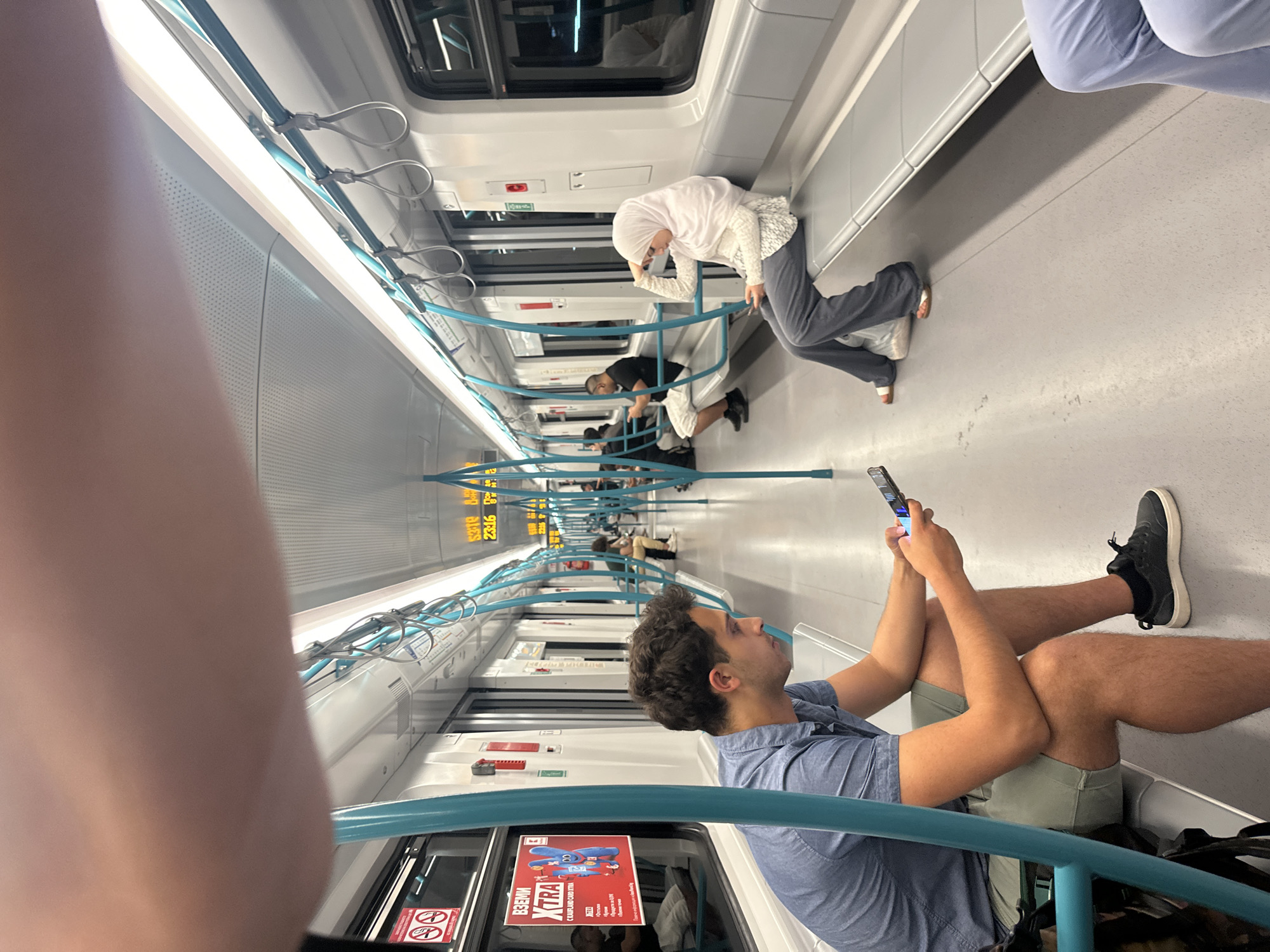 The image depicts the interior of a modern, light-colored subway train carriage with teal-colored vertical poles and grab handles. The floor is gray and there are rows of gray seats along the sides. In the foreground, a man in a blue short-sleeved shirt and gray shorts is seated, looking down at his mobile phone. To his right, further down the carriage, a woman wearing a white hijab and a white and gray patterned top with gray pants is seated, also looking at her phone and resting her head on her hand. Several other passengers are visible further inside the carriage, some seated and some standing. An advertisement poster featuring a cartoon character is visible on the window to the left. Digital displays showing times in yellow are visible above the windows. The overall impression is of a typical public transport environment. The context of Sofia, Bulgaria, implies this is likely a metro carriage within the city's transit system.