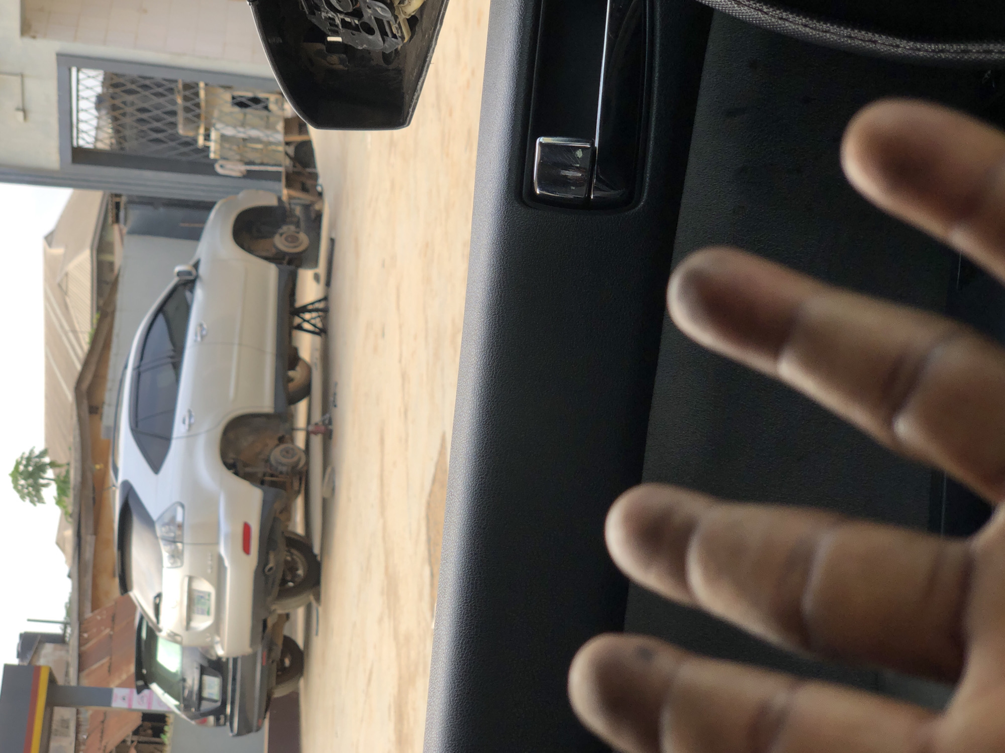 The image depicts a street scene in Onibudo, Nigeria, focusing on two cars undergoing maintenance in what appears to be a repair bay. In the foreground, the interior of a vehicle is visible, with a person's hand, smudged with dirt, obscuring part of the view. The hand is positioned close to the camera, suggesting the photo was taken from inside this vehicle.
Dominating the mid-ground are two SUVs. A silver SUV is centrally positioned, with all four wheels removed and the vehicle supported by jack stands. Its undercarriage is exposed, indicating mechanical work is in progress. To the left of the silver SUV is a darker colored SUV, partially visible, also appearing to be undergoing some form of service.
The background reveals elements of the urban environment. Buildings with corrugated iron roofs are visible, along with what looks like a workshop or garage structure with a metal gate. A street or paved area serves as the ground surface where the vehicles are located. The overall impression is of an active automotive service area within the Nigerian city.