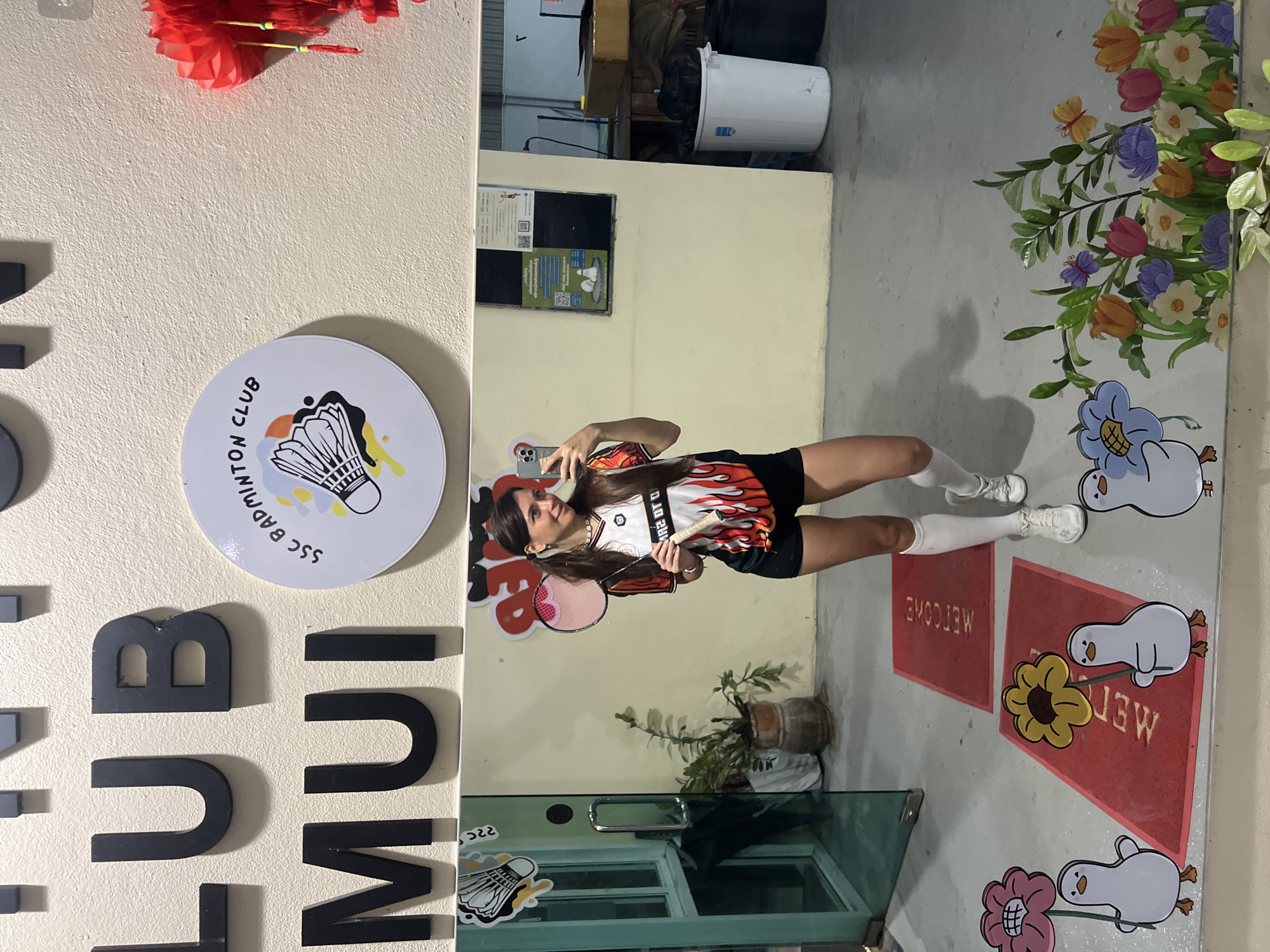 A young woman is taking a mirror selfie in what appears to be a badminton club or recreational sports facility. She is dressed in a red and white flame-patterned sports jersey, black shorts, and white socks. She holds her phone in her left hand and a badminton racket in her right.
The reflection in the mirror shows a room with a tiled floor adorned with cartoonish decorations, including cartoon flowers, butterflies, and duck characters. There are also red mats with "WELCOME" printed on them. The wall behind her is decorated with the letters "CLUB" in large black capital letters and a round sticker with the words "SSC Badminton Club" and a shuttlecock design.
A potted plant sits near the bottom of the mirror, and a door with glass panels is also visible. In the background, there are glimpses of a more utilitarian setting with a white bucket, storage containers, and various signs on the wall.