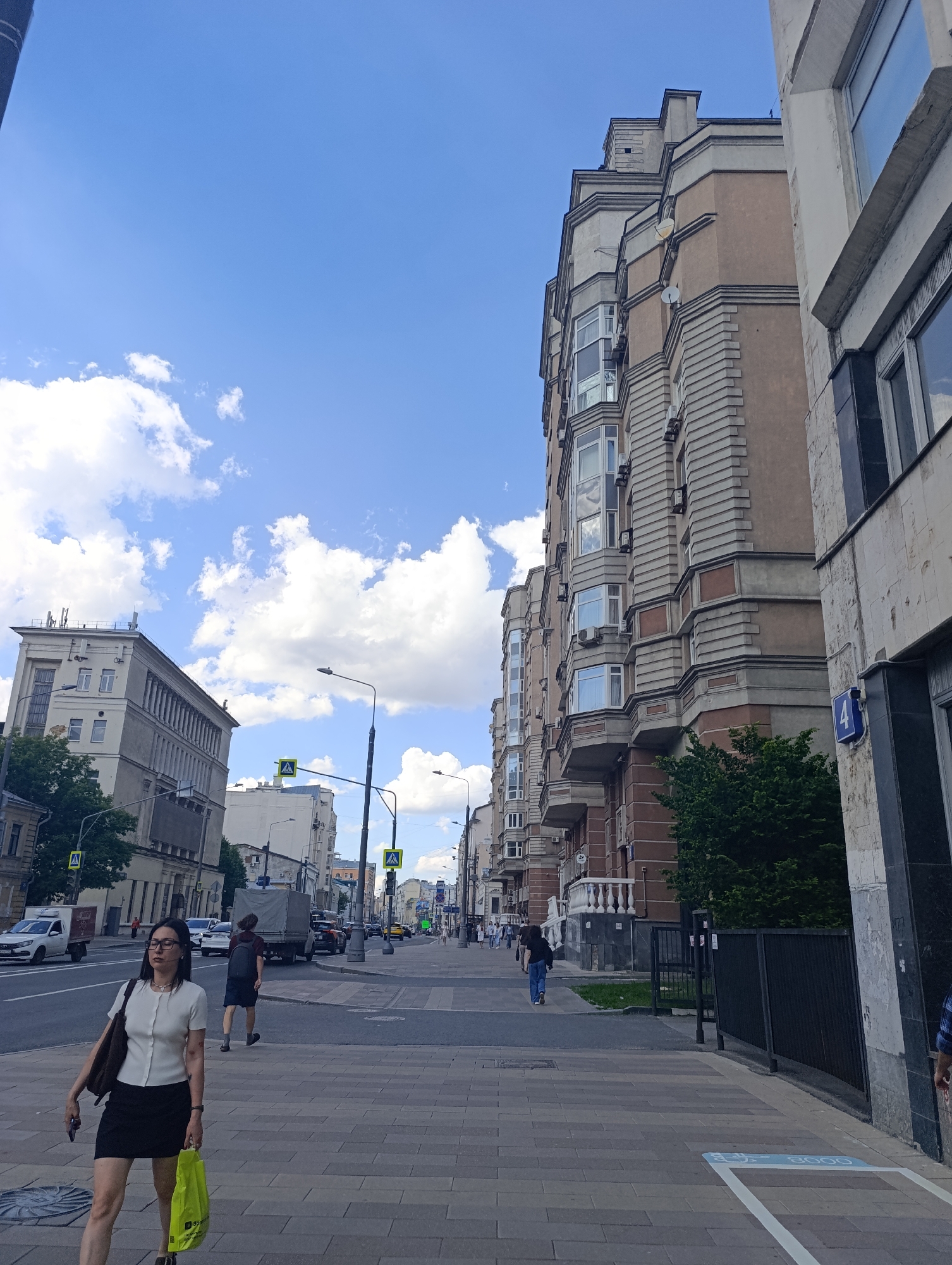 The image presents a daytime street scene in Moscow, Russia, under a clear blue sky with scattered white cumulus clouds. Bright daylight illuminates the urban environment, suggesting it's mid-morning or afternoon.
In the foreground, a woman wearing glasses, a white short-sleeved top, and a black skirt walks on a patterned paved sidewalk, heading towards the left. She carries a dark shoulder bag and a bright yellow shopping bag, which partially shows "3g" text. She holds a mobile phone in her left hand. Further down the street, a man with a backpack walks away from the camera on the road, near parked and moving vehicles, including a white van and a dark truck. Other pedestrians are visible in the distance.
The street is lined with multi-story buildings. On the right, a prominent building with intricate architectural details, including bay windows, cornices, and textured panels in shades of beige and brown, dominates the frame. A blue square sign with the white number "4" is visible on its facade. Several small satellite dishes are mounted on this building. Further along the street on the left, another large building with a lighter facade, featuring columns and numerous windows, stands out. Streetlights and yellow pedestrian crossing signs with an "A" symbol are present along the road.
On the sidewalk in the bottom right, a partially visible blue painted square outline contains an icon and the number "8000", possibly indicating a marker for a service or distance. The scene captures everyday city life with people commuting and vehicles in transit.