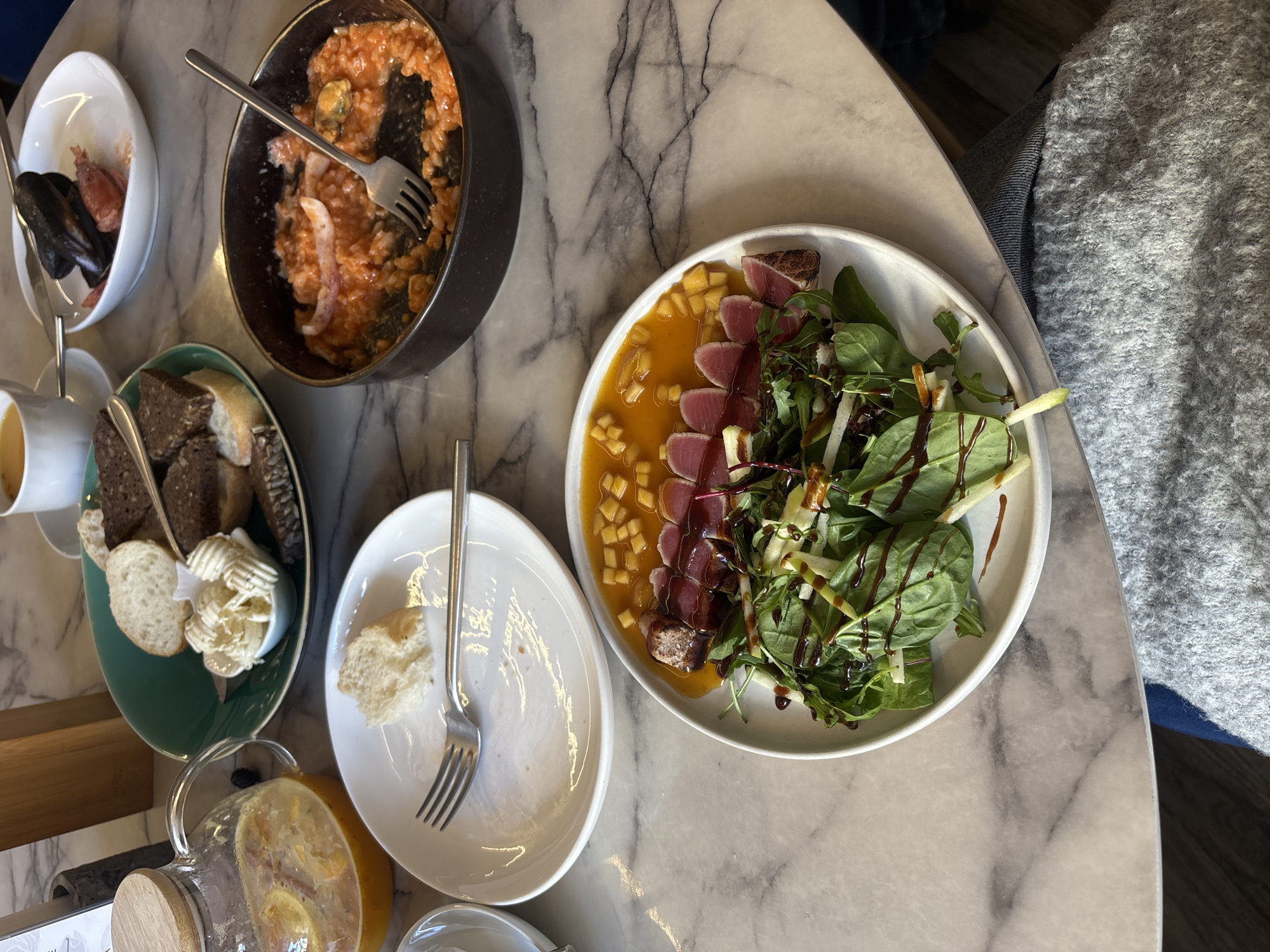 The image depicts a table with several dishes of food. There is a salad with sliced tuna, greens, and dressing; a dish of what appears to be seafood risotto with mussels; a bowl of bread with a small bowl of butter; a small bowl of mussels; and a teapot of juice. A fork rests on a mostly empty plate. The table appears to be marble. A person wearing a gray sweater is partially visible on the right. The location cannot be determined from the image.
