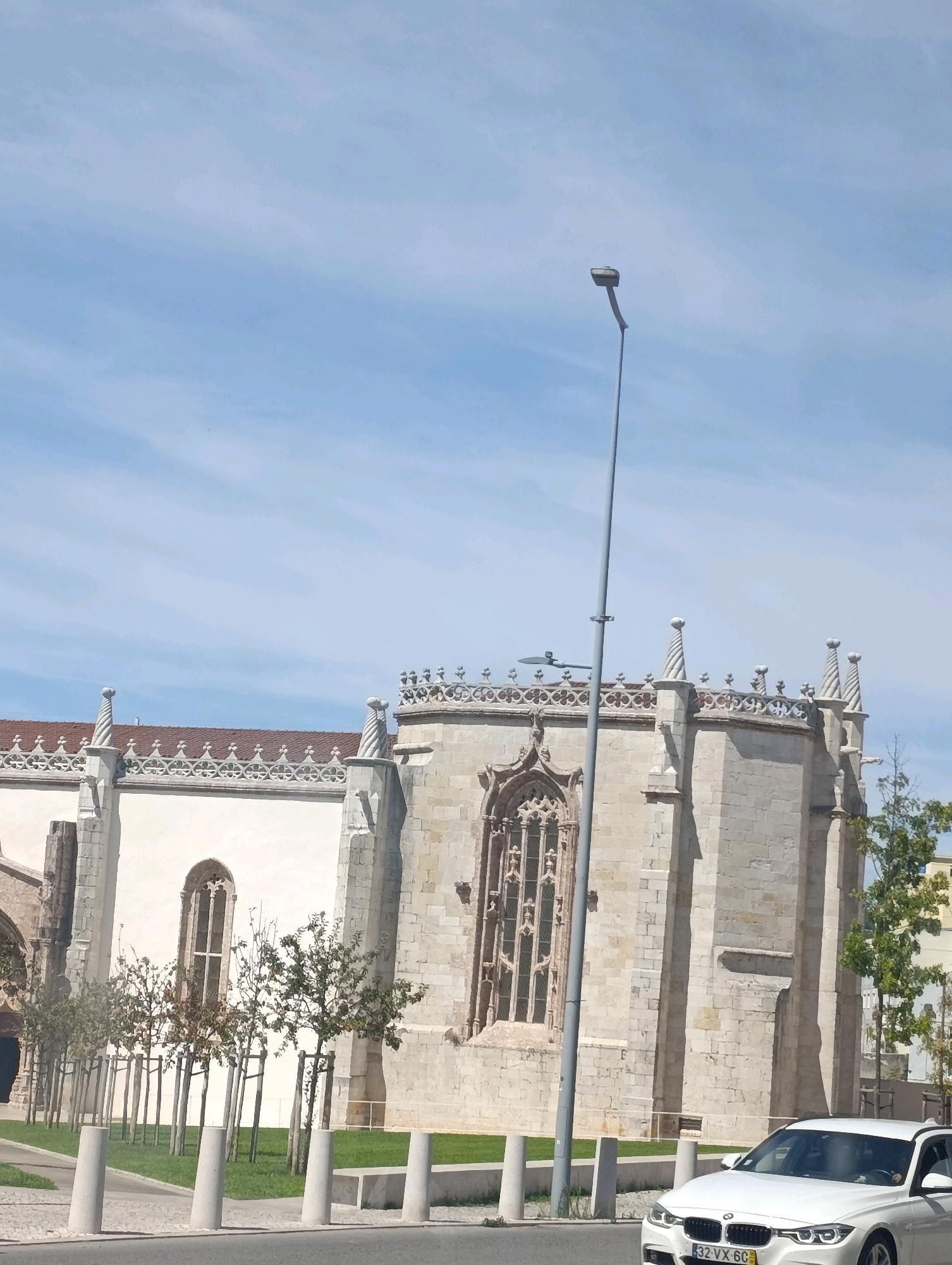 The image shows a white BMW parked in front of a large, light-colored stone building with ornate architectural details and a red tiled roof. The building appears to be a church or a historical monument. There are several trees in front of the building, some of which are bare. The sky is blue with some scattered clouds. A lamppost stands in the foreground to the left of the building. The license plate of the car reads "32 VX 6C". This scene is located in Setúbal, Portugal, specifically in front of the Convento de Jesus de Setúbal, a former convent and a National Monument.