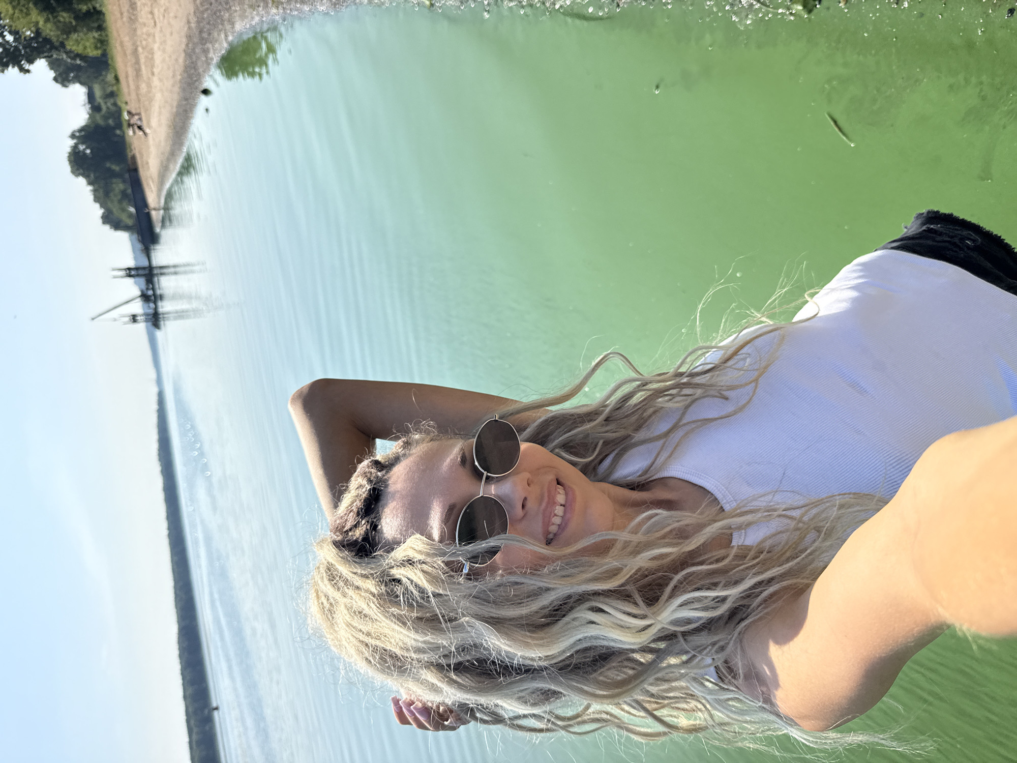 A blonde woman with long, wavy hair and sunglasses is taking a selfie on a wooden pier. She is wearing a white tank top and black shorts. The water is a murky green color, and the sky is clear and blue. In the background, there is a beach with trees and a crane. The location is the city of Rusanovo, Russia.