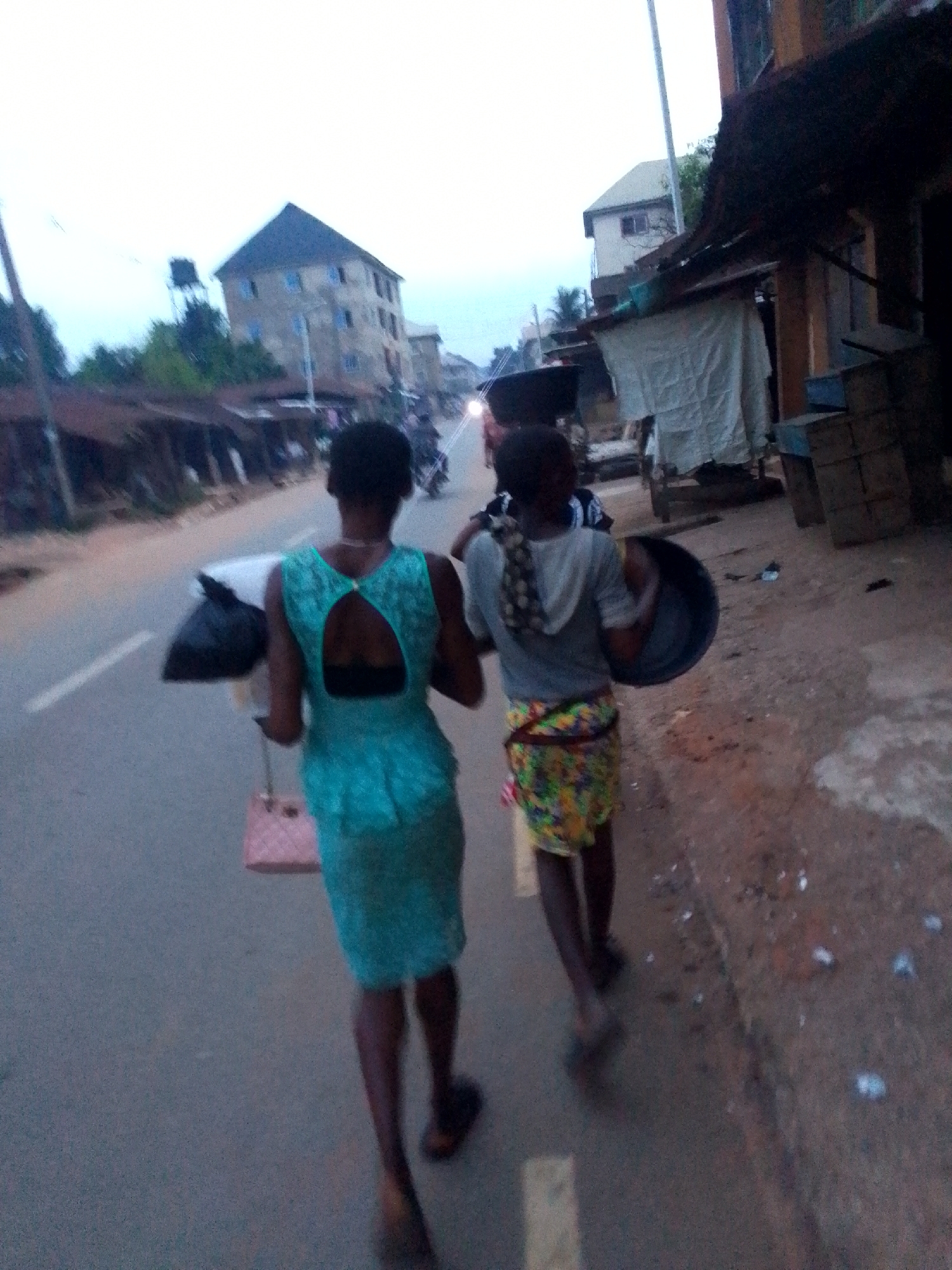 The image depicts a street scene in Orlu, Nigeria, likely during dusk or dawn due to the dim, diffused natural light under an overcast sky. Two females are walking away from the camera along a paved road.
The woman in the foreground, on the left, has short dark hair and wears a sleeveless, teal-colored dress featuring a triangular cutout design on the back. She is carrying a black plastic bag and a pink quilted handbag, which appears to be attached to a small, wheeled frame. She wears dark sandals.
The second woman, slightly behind and to the right, also has short dark hair and a patterned cloth tied around her neck or head. She is dressed in a light grey hooded top and a vibrant, multi-colored patterned skirt. She carries a large, dark, shallow bowl or pan in her left hand.
The road surface is asphalt with a faint white broken line. To the left of the road, there are some sparse trees and distant multi-story buildings, one featuring a dark roof and a visible water tank on a stand. To the right, the roadside transitions into a dirt path lined with makeshift market stalls or shops, some covered with white tarpaulins, and stacked wooden crates. A bright light, possibly from a distant vehicle, is visible further down the road. No legible text is visible in the frame, and the emotions of the subjects cannot be discerned as their backs are to the camera.