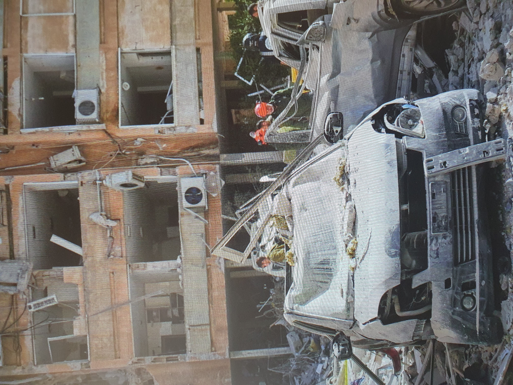 The image depicts the aftermath of an explosion in a residential building in Tashkent, Uzbekistan. The building, a multi-story structure with visible damage including blown-out windows and collapsed sections, appears to be made of concrete and brick. In the foreground, two vehicles are heavily damaged, covered in debris. One car, a light-colored hatchback, is positioned front and center, its hood significantly crumpled and its front end destroyed. Behind it, a dark-colored SUV is also visible, its side heavily impacted.
Several individuals, appearing to be emergency responders or rescue personnel, are present at the scene. Some are wearing orange vests and helmets, suggesting they are involved in search and rescue operations. One person, wearing a helmet and green uniform, is visible near the damaged car. The environment is filled with rubble, concrete fragments, and twisted metal, indicative of a severe blast. Wires and debris are scattered around the scene, adding to the chaotic nature of the aftermath. The overall impression is one of significant destruction and ongoing rescue efforts.