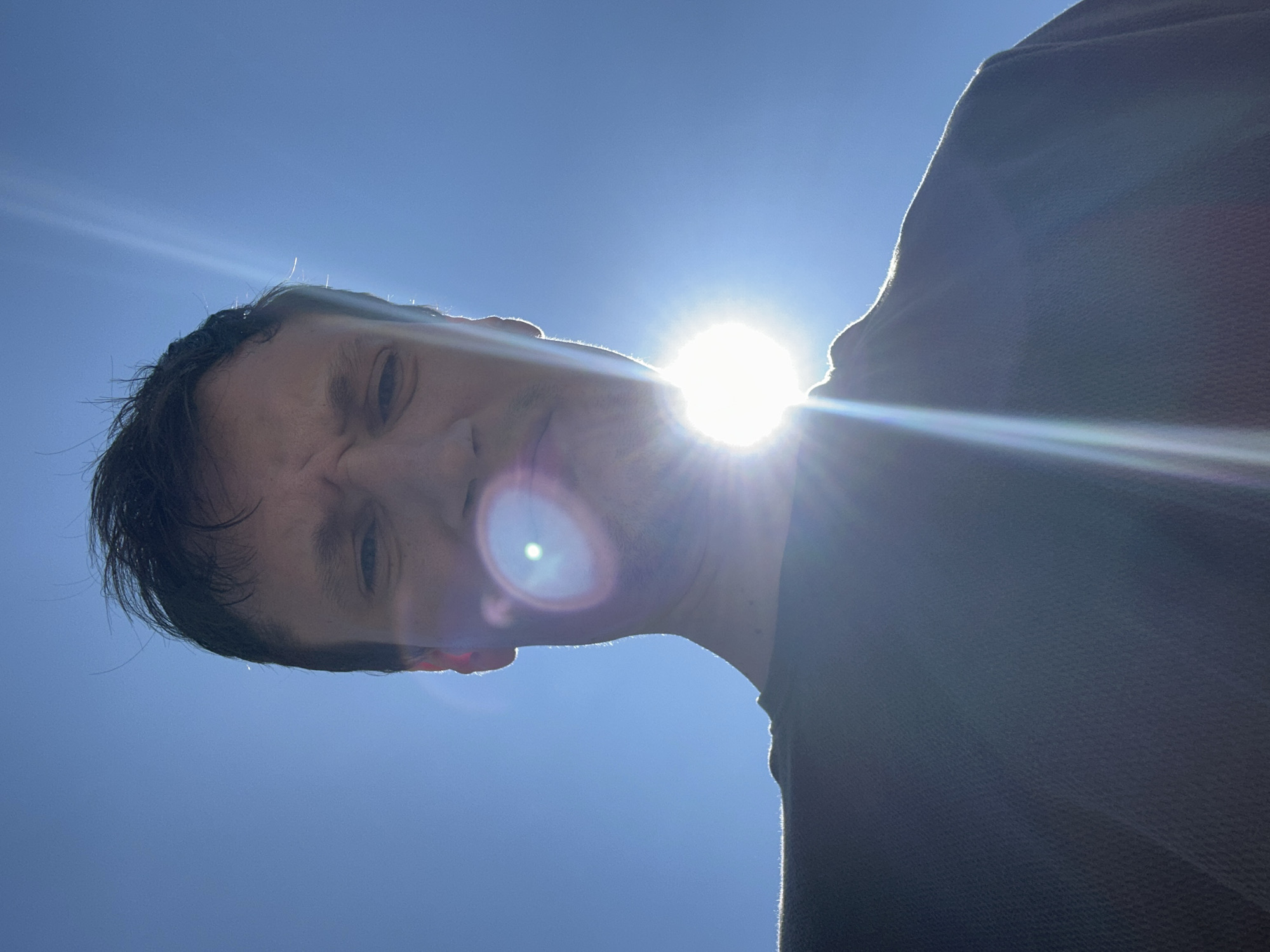 A man is captured in a low-angle, outdoor shot, with the bright sun positioned directly behind his head, creating lens flare and a silhouette effect. The sky is a clear, deep blue. The man's face is partially obscured by the intense sunlight, but his brow is furrowed, and his mouth is slightly downturned, suggesting a pained or critical expression. He is wearing a dark crew-neck shirt. The image does not contain enough visual information to determine the specific location in Shatava, Ukraine.