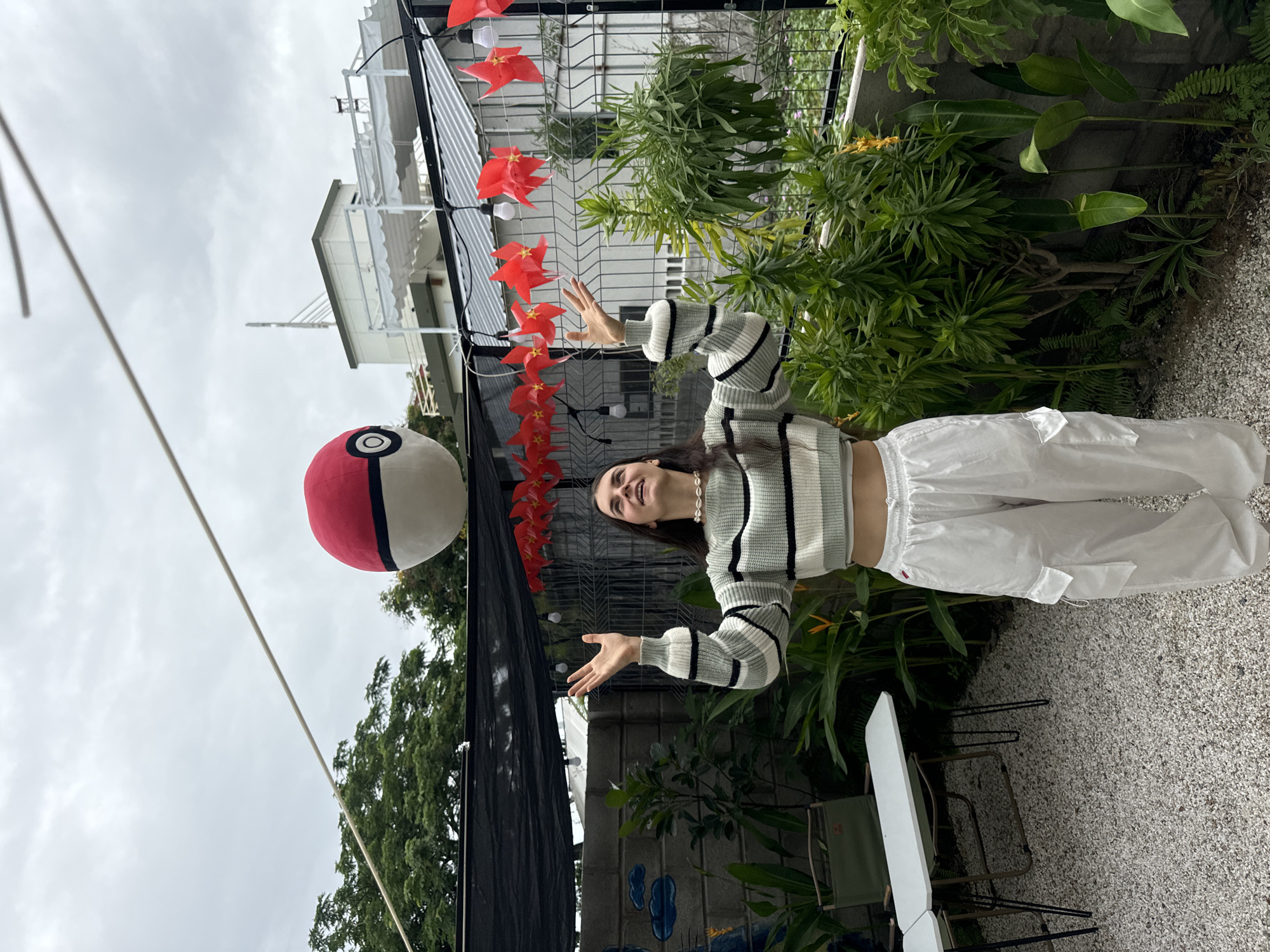 A young woman stands in what appears to be an outdoor cafe or garden setting in Phu Thuy Ward, Vietnam. She is positioned between a large, inflated Poké Ball on the left and hanging red decorations overhead. The woman is wearing a cropped, striped sweater and loose, white pants. Her arms are raised with palms facing upwards, as if gesturing towards the Poké Ball or the decorations. The background includes greenery, a building with a corrugated metal roof, and a gray sky. Chairs and tables are visible on the ground, which appears to be covered in gravel.