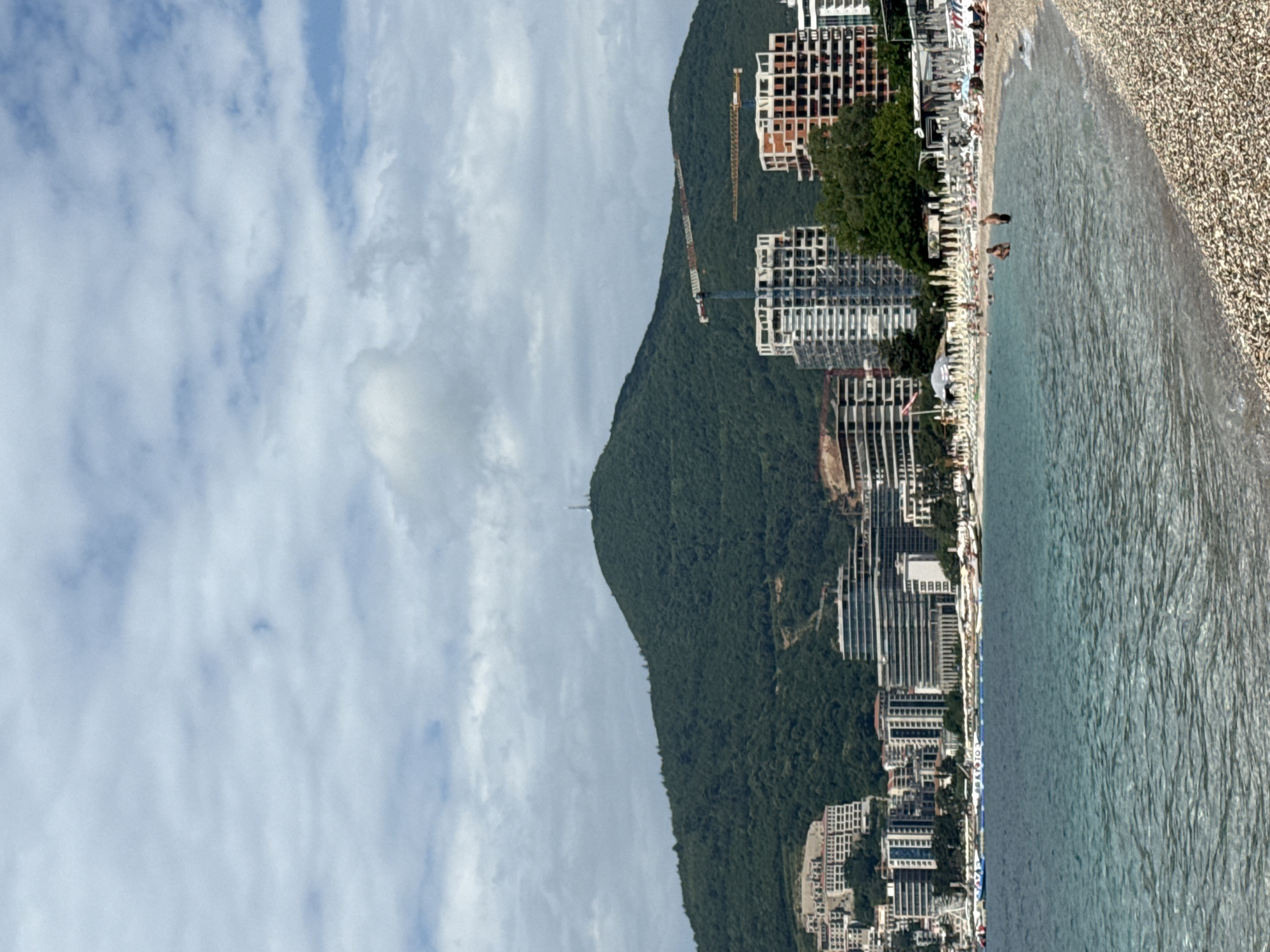 The image shows a vertical view of a coastal scene in Budva, Montenegro. A pebble beach borders a body of water with small waves. A dense hillside covered in green trees rises behind the beach, dotted with buildings and construction cranes. Tall structures and buildings line the shoreline. The sky has light, scattered clouds. Some people are visible along the beach.