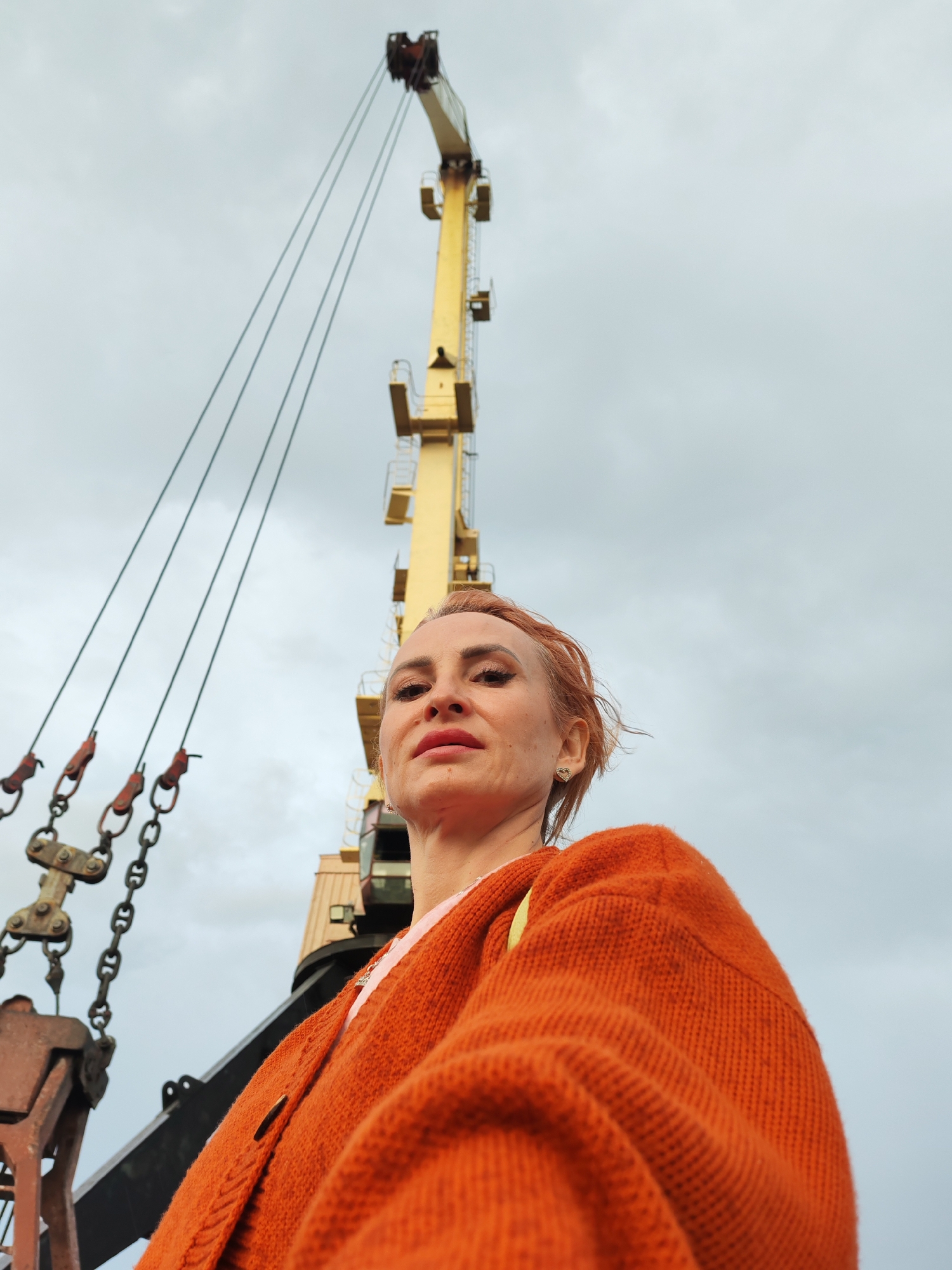 An upward-angle, medium shot captures a fair-skinned woman with reddish-brown hair styled in a messy bun, looking slightly upwards and to the left. She is wearing an orange knitted cardigan over a pink garment and a gold heart-shaped earring. Her makeup includes winged eyeliner and red lipstick. The background is dominated by the cloudy sky and a large, yellow industrial crane. The crane's structure, including its boom and cables, extends upwards and diagonally across the frame. Part of a rusty industrial mechanism with chains is visible in the lower left quadrant. The overall setting appears to be a port or industrial area, possibly in Shalsky, Russia.